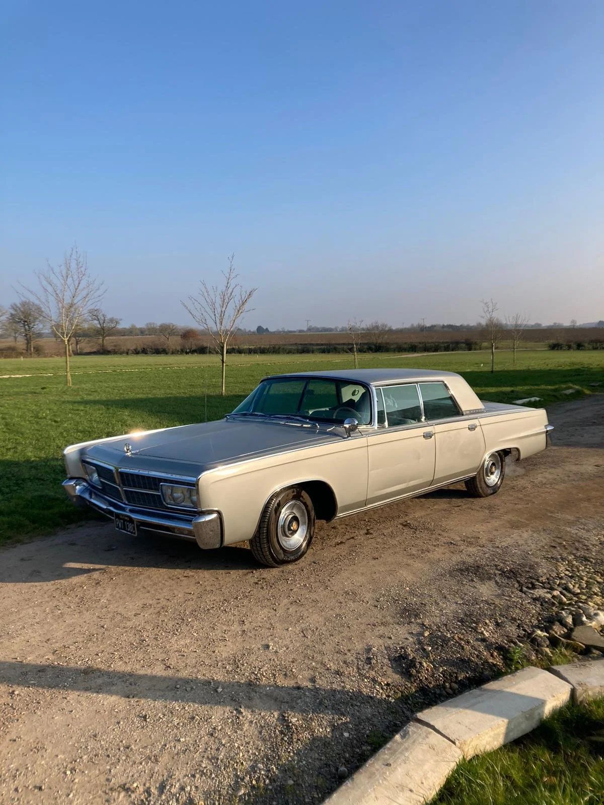 Vintage white sedan parked on dirt beside a grassy field with a few leafless trees and a clear blue sky.