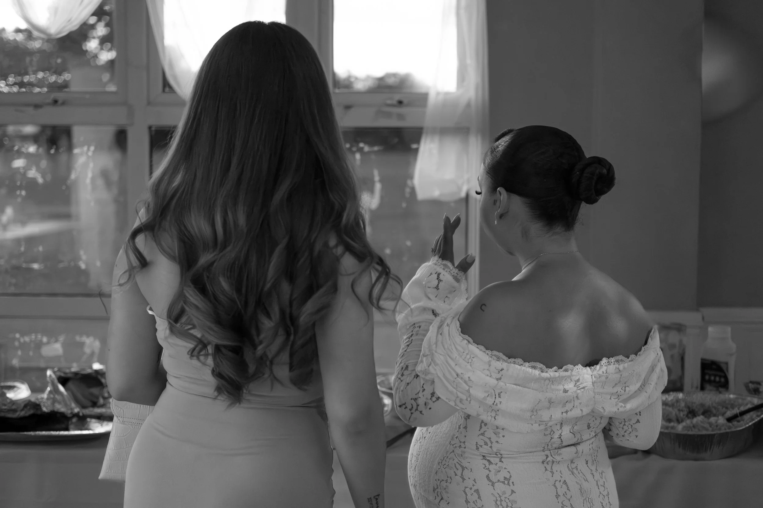 Two women standing by a window, engaged in conversation, with a table of food behind them. The woman on the left has long wavy hair and is wearing a sleeveless dress, while the woman on the right has her hair in a bun and is wearing an off-the-should