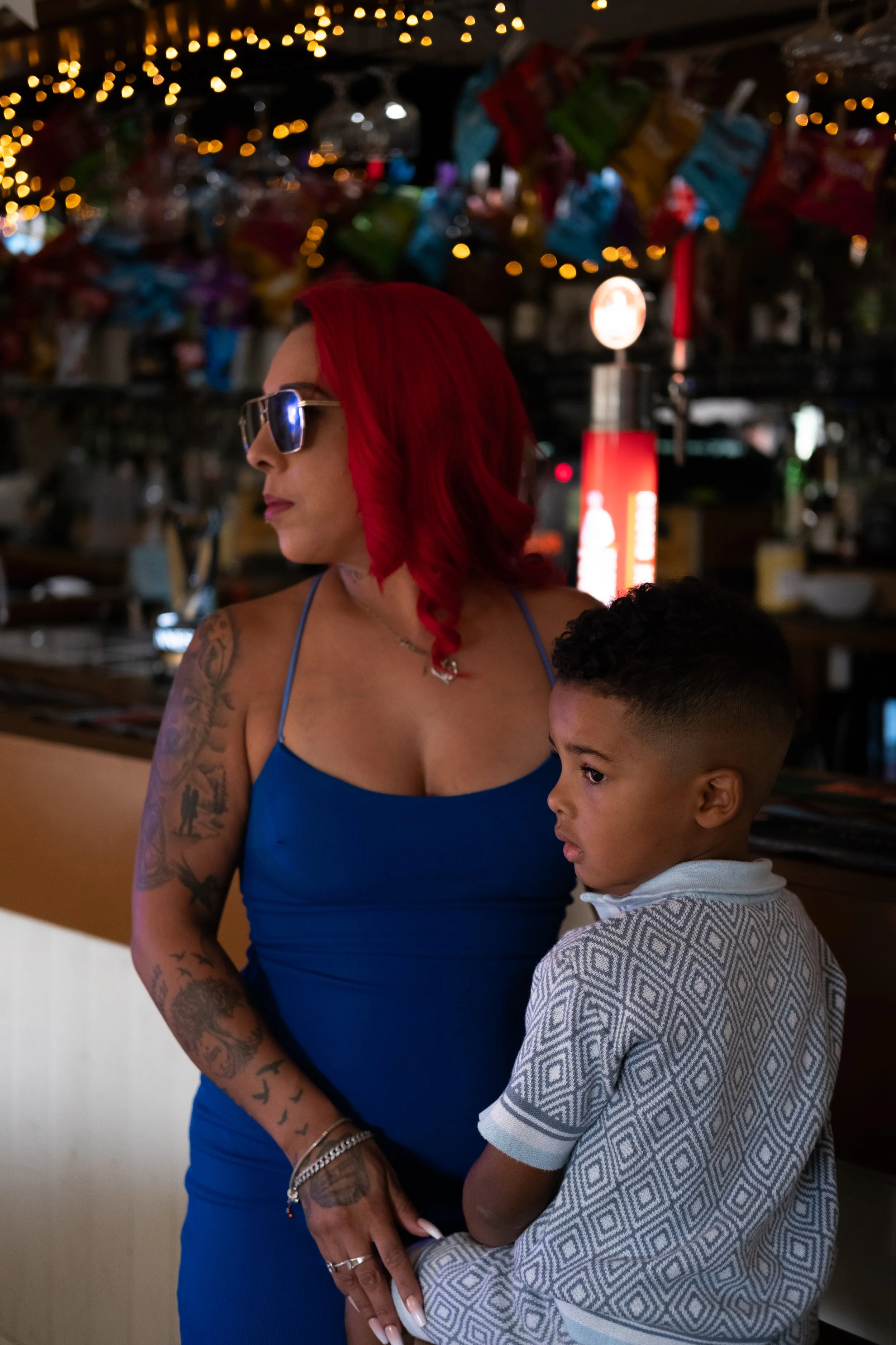 A woman with red hair, sunglasses, and tattoos wearing a blue dress stands next to a young boy with short curly hair in a patterned shirt inside a dimly lit bar or restaurant decorated with string lights and colorful items hanging from the ceiling.