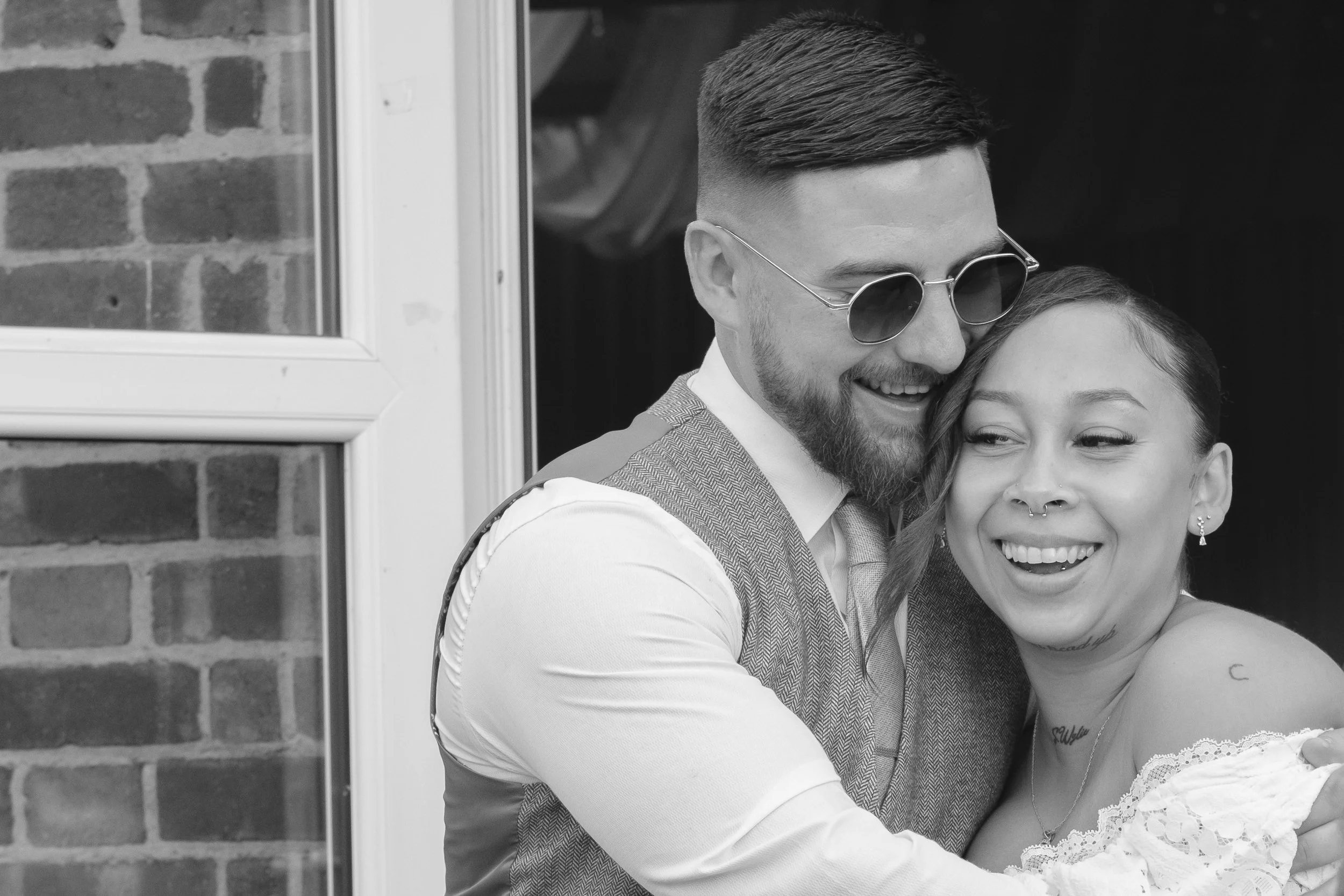 A black and white photo of a happy couple smiling and hugging each other, standing outside near a brick wall and a window.