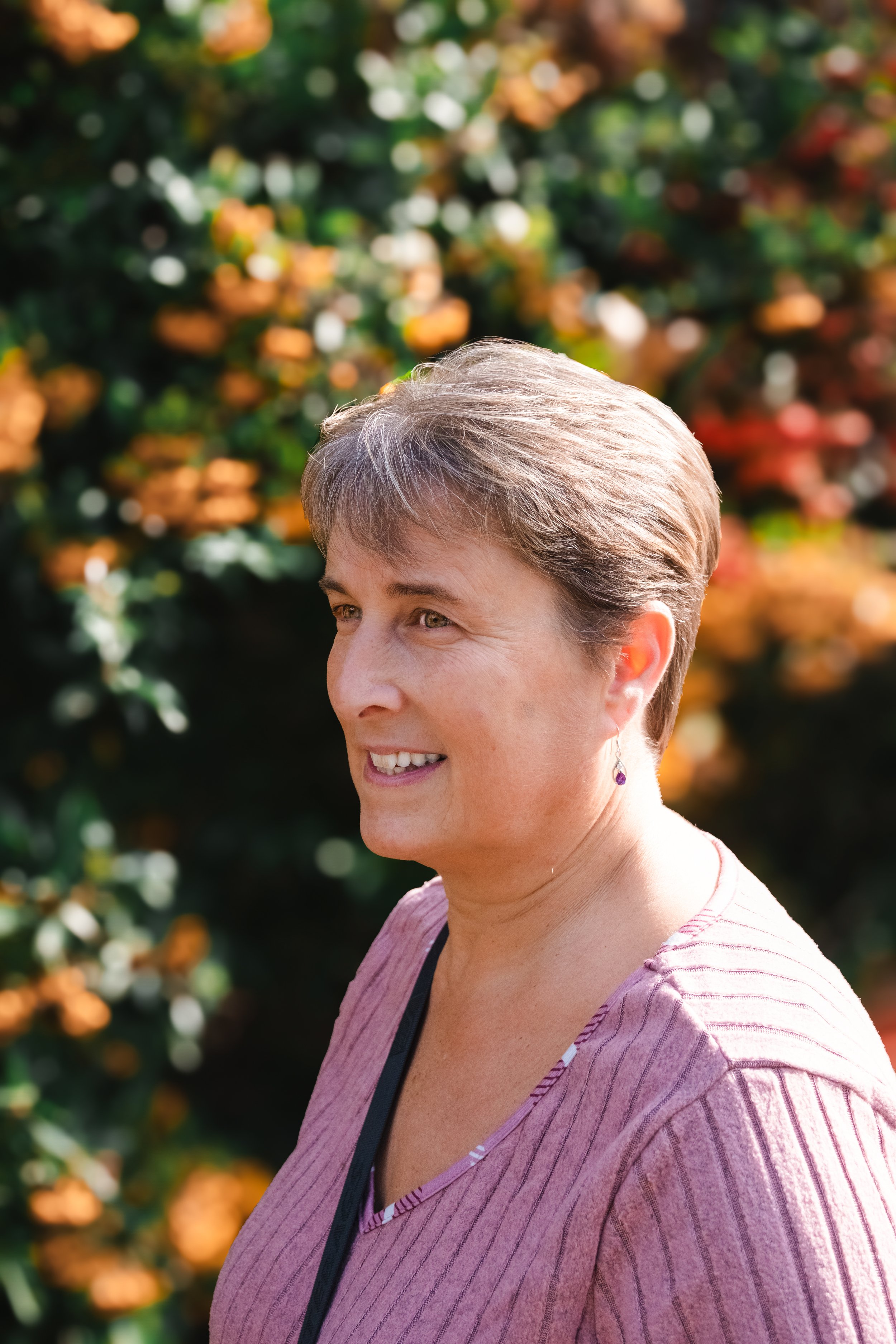 Portrait of a smiling woman with short, gray hair outdoors, with blurred colorful foliage in the background.