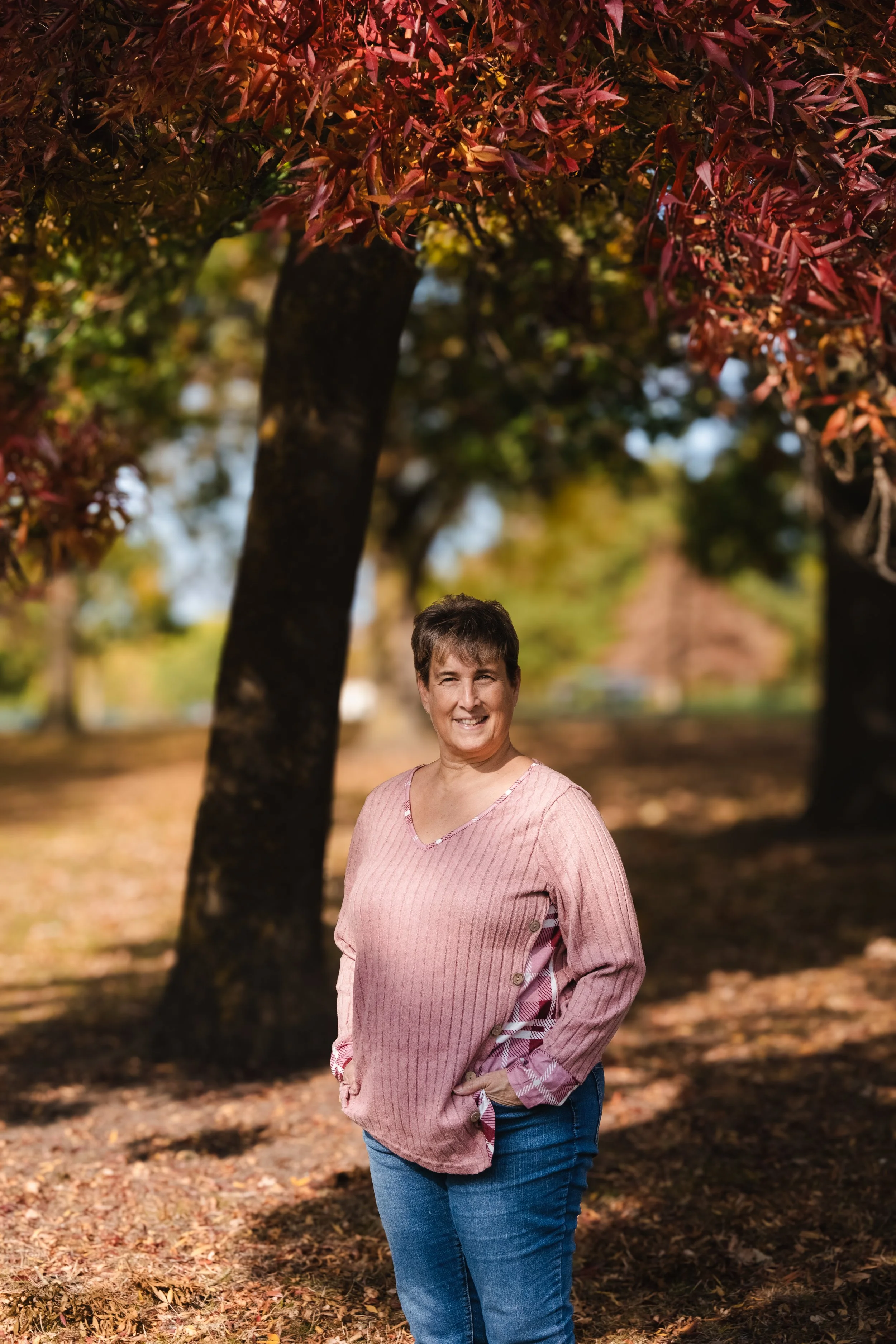 A woman standing outdoors under fall trees with red and orange leaves, smiling with hands in pockets, wearing a pink sweater and blue jeans.