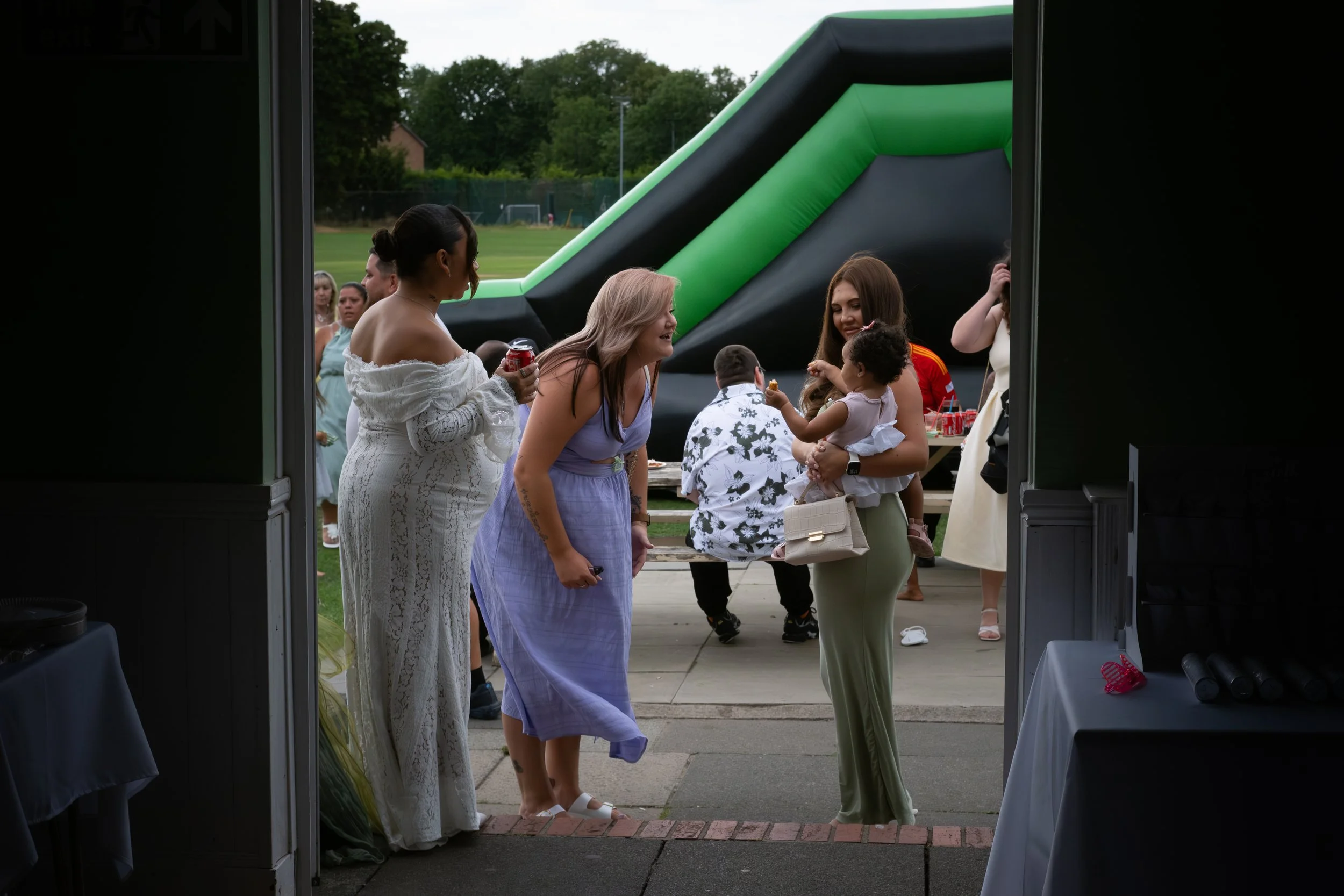 People gathered outside a building during a celebration, with some engaging and smiling at each other, and a large inflatable slide in the background, on a grassy field with trees.