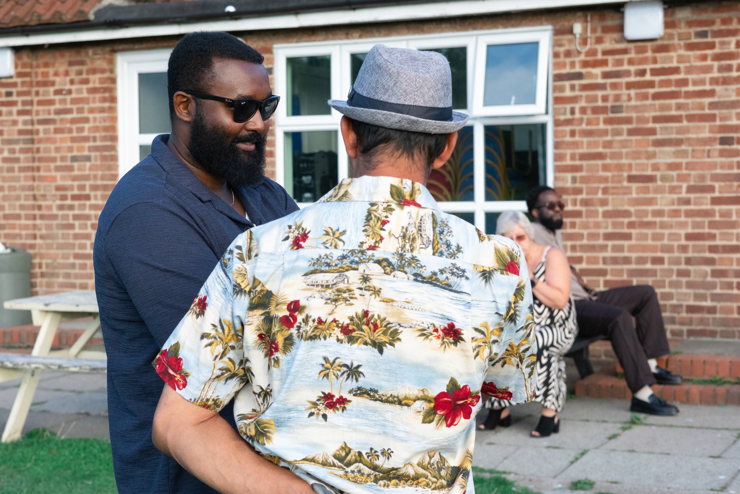 Two men are engaged in conversation outdoors in front of a brick building, with two women sitting on a bench in the background.