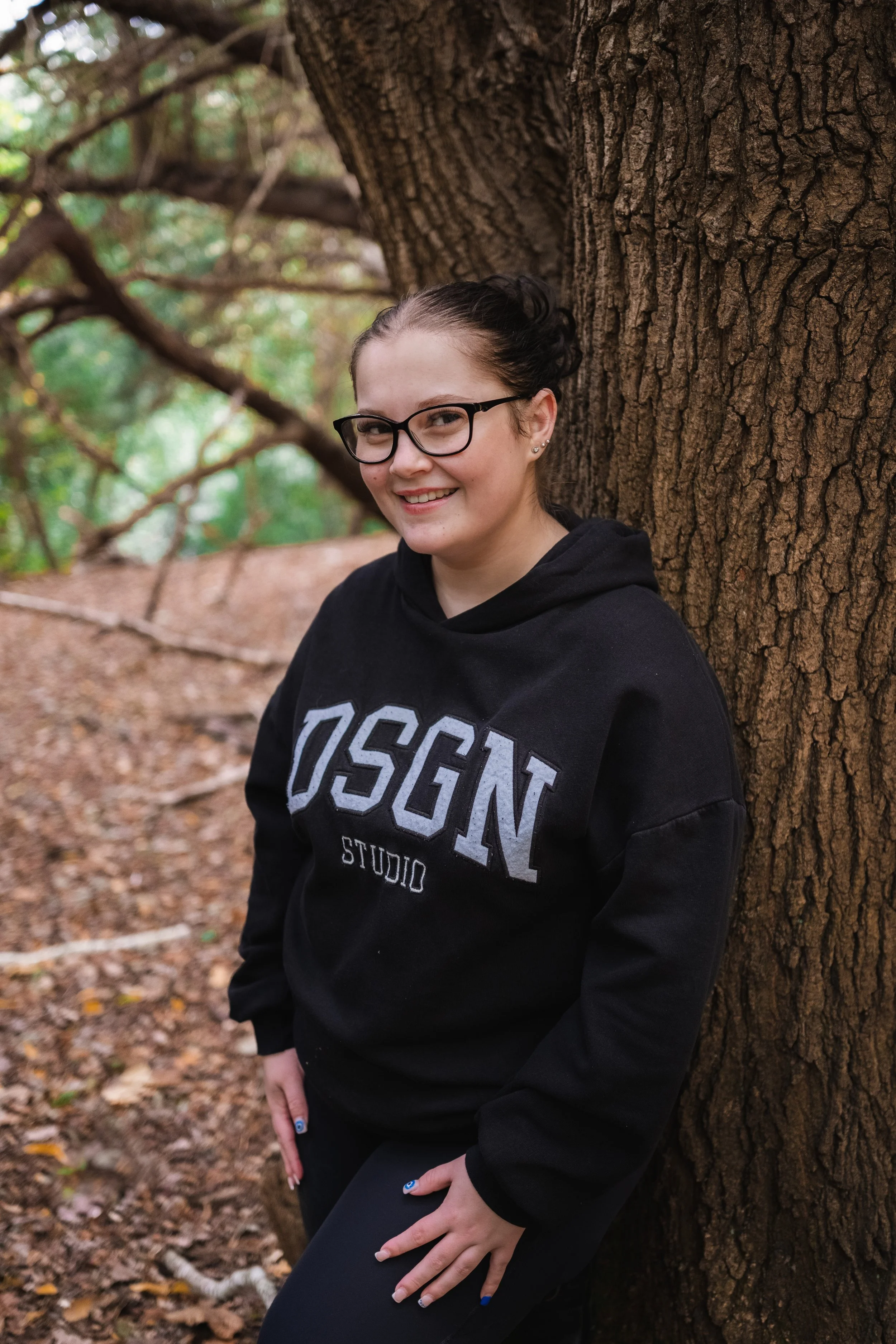 A young woman with glasses and dark hair tied back wearing a black hoodie with 'OSGN STUDIO' printed on it, leaning against a tree in a wooded area with fallen leaves on the ground.