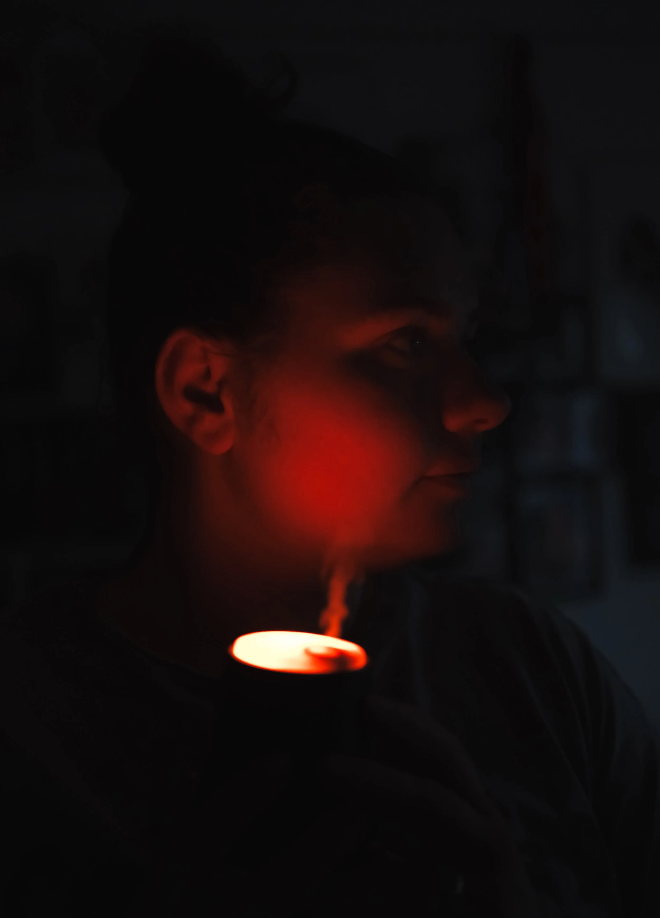 A woman in a dark room exhaling vapor from a vape or e-cigarette, with a faint glow of red light illuminating her face.