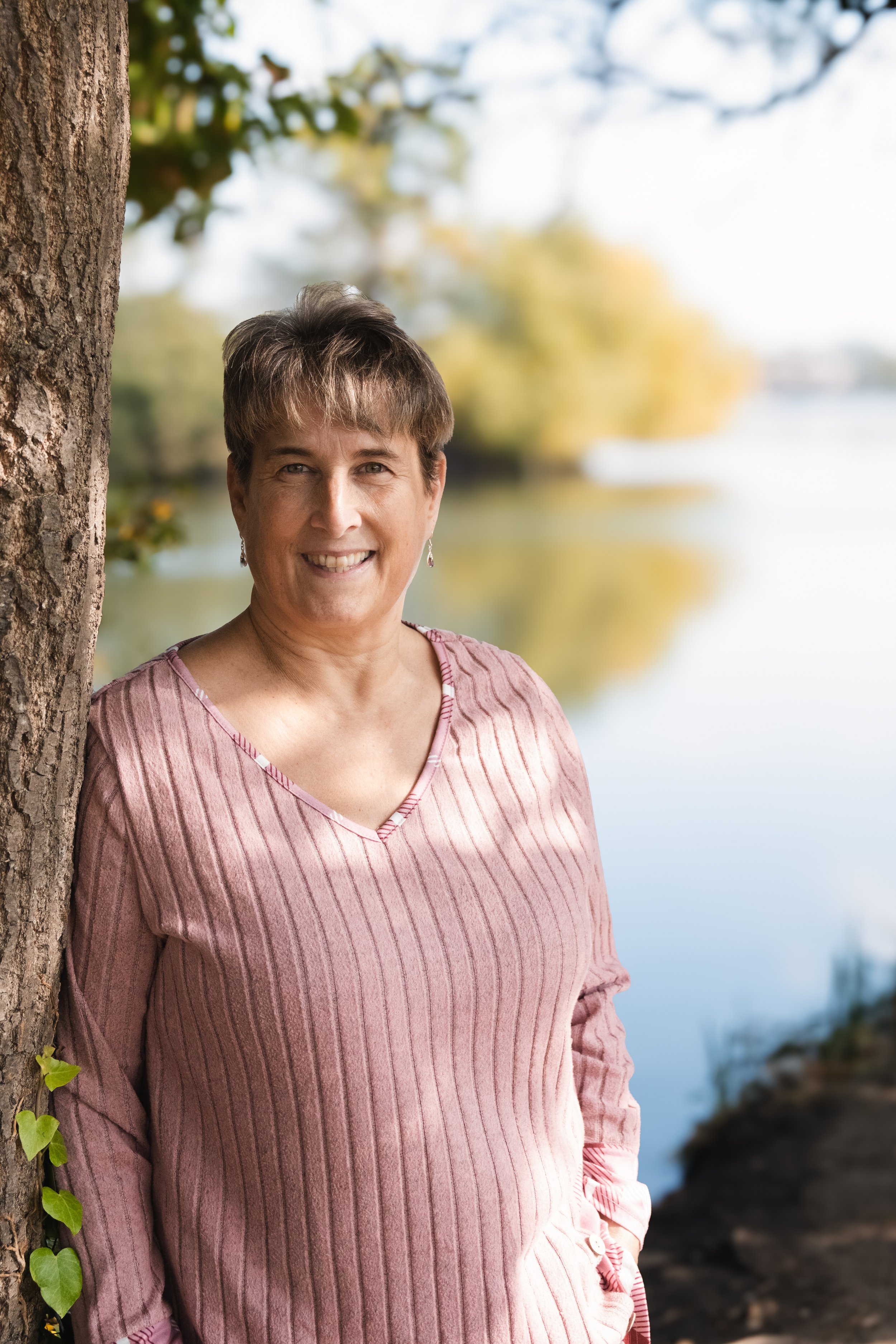 A woman with short brown hair wearing a pink striped shirt, standing outdoors near a tree by a body of water with trees and sky in the background.