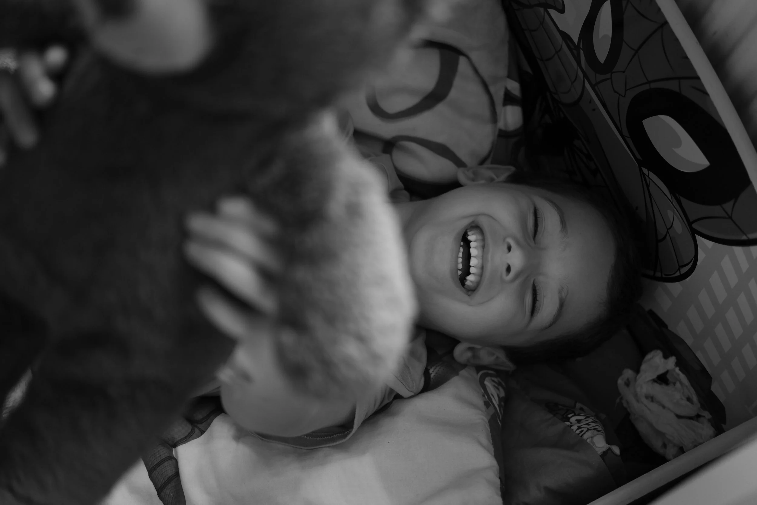 A young girl lying in bed, smiling and laughing as she interacts with a stuffed animal, in a child's room with patterned bedding.