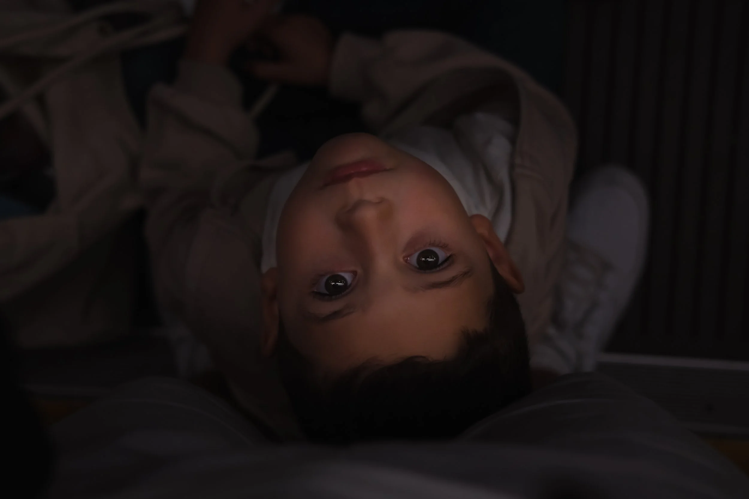 A young boy lying on his back, looking up at the camera with wide eyes, in a dark room.