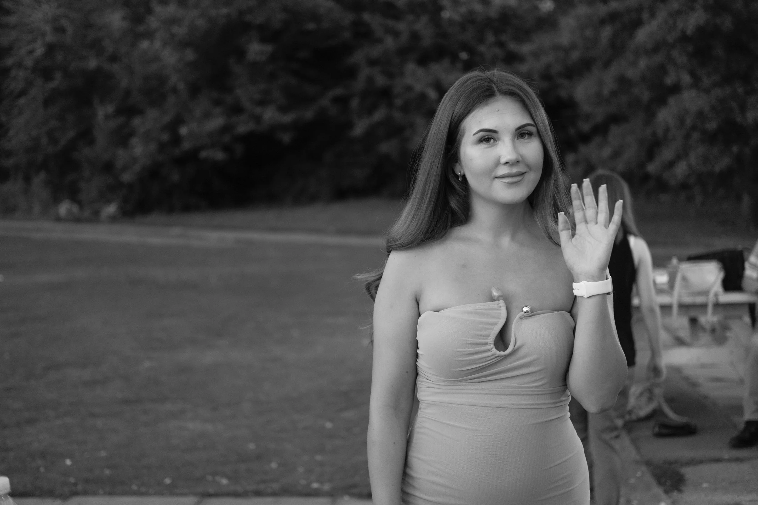 A woman with long hair waving at camera, wearing a strapless dress, outdoors in a park or garden setting.