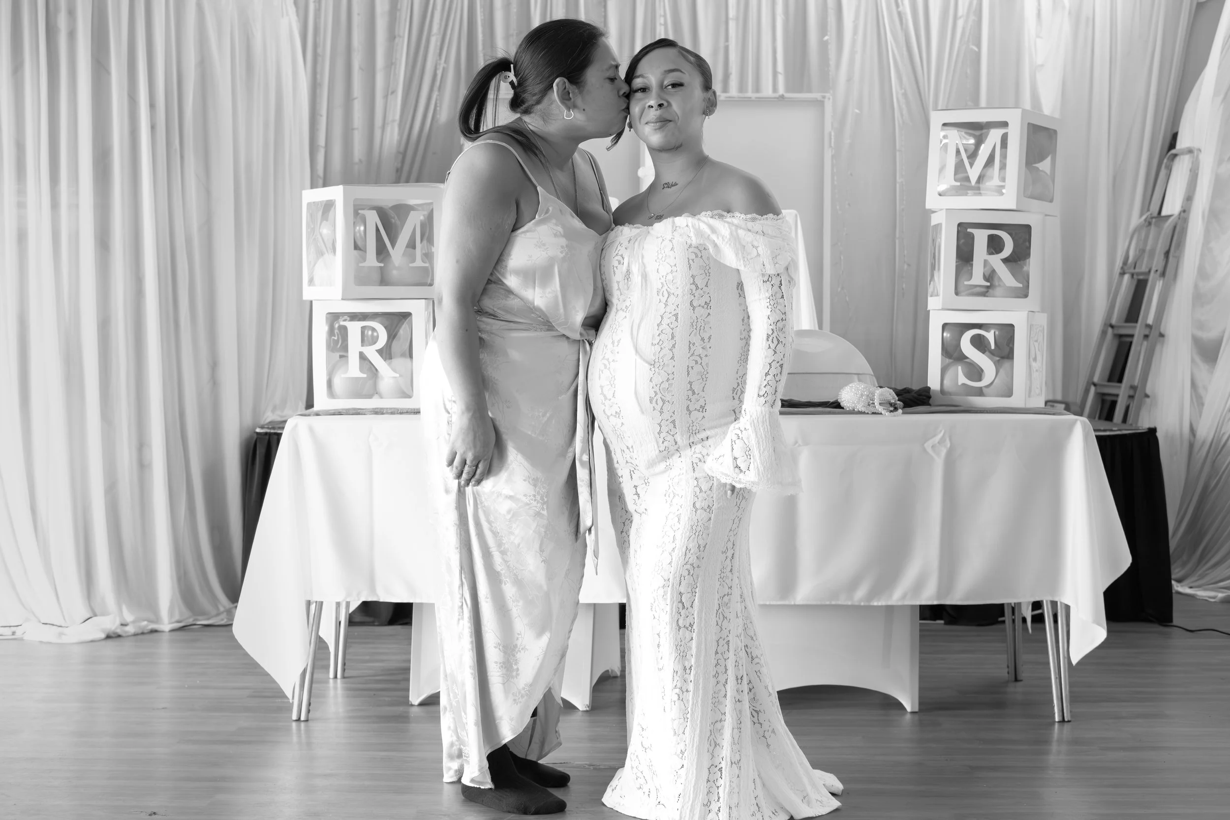Two women, one kissing the other's cheek, dressed in satin and lace gowns, in front of a table with decorative blocks spelling 'MORS'.