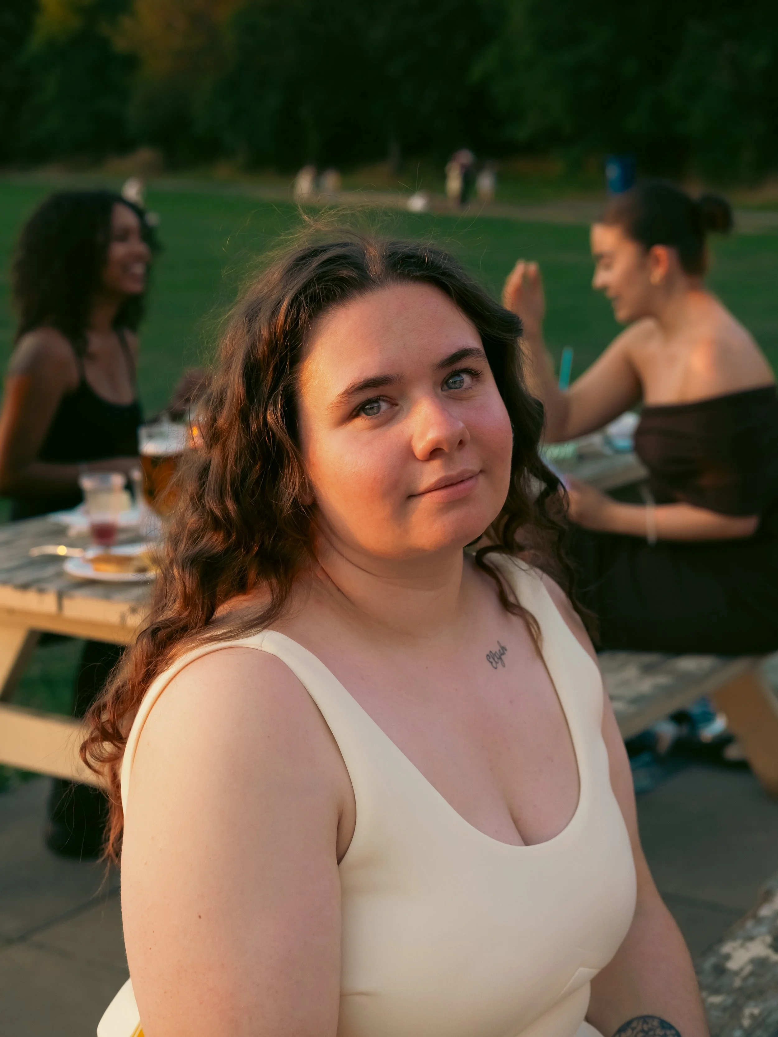 A woman with long, curly brown hair wearing a cream-colored sleeveless top, sitting outdoors during sunset with a tattoo on her chest that says "hope." In the background, two women are laughing and talking at a picnic table with food and drinks, in a