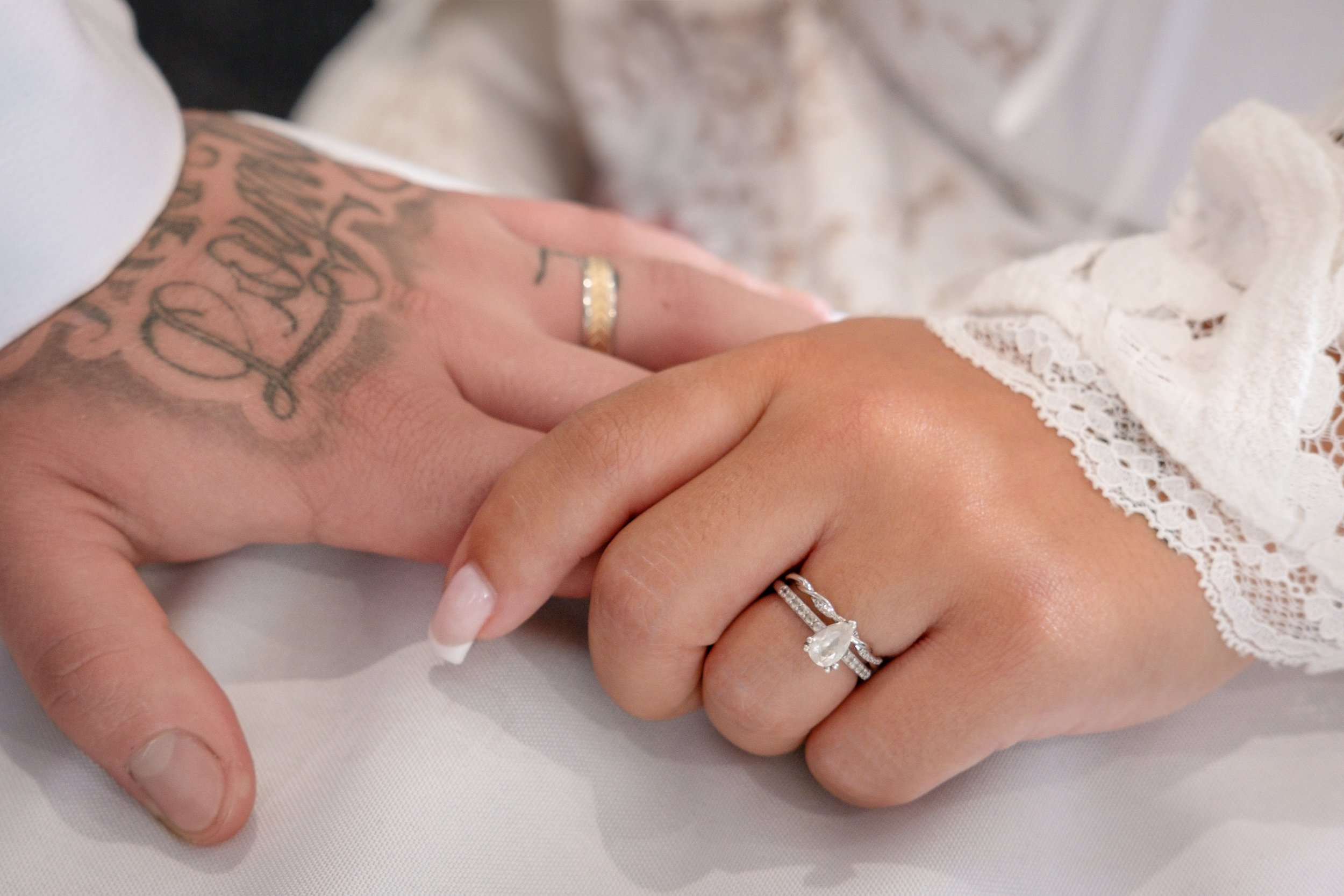 Close-up of a couple's hands showing wedding rings, one with a wedding band and a diamond engagement ring, with lace on the woman's sleeve and tattoos on the man's hand.