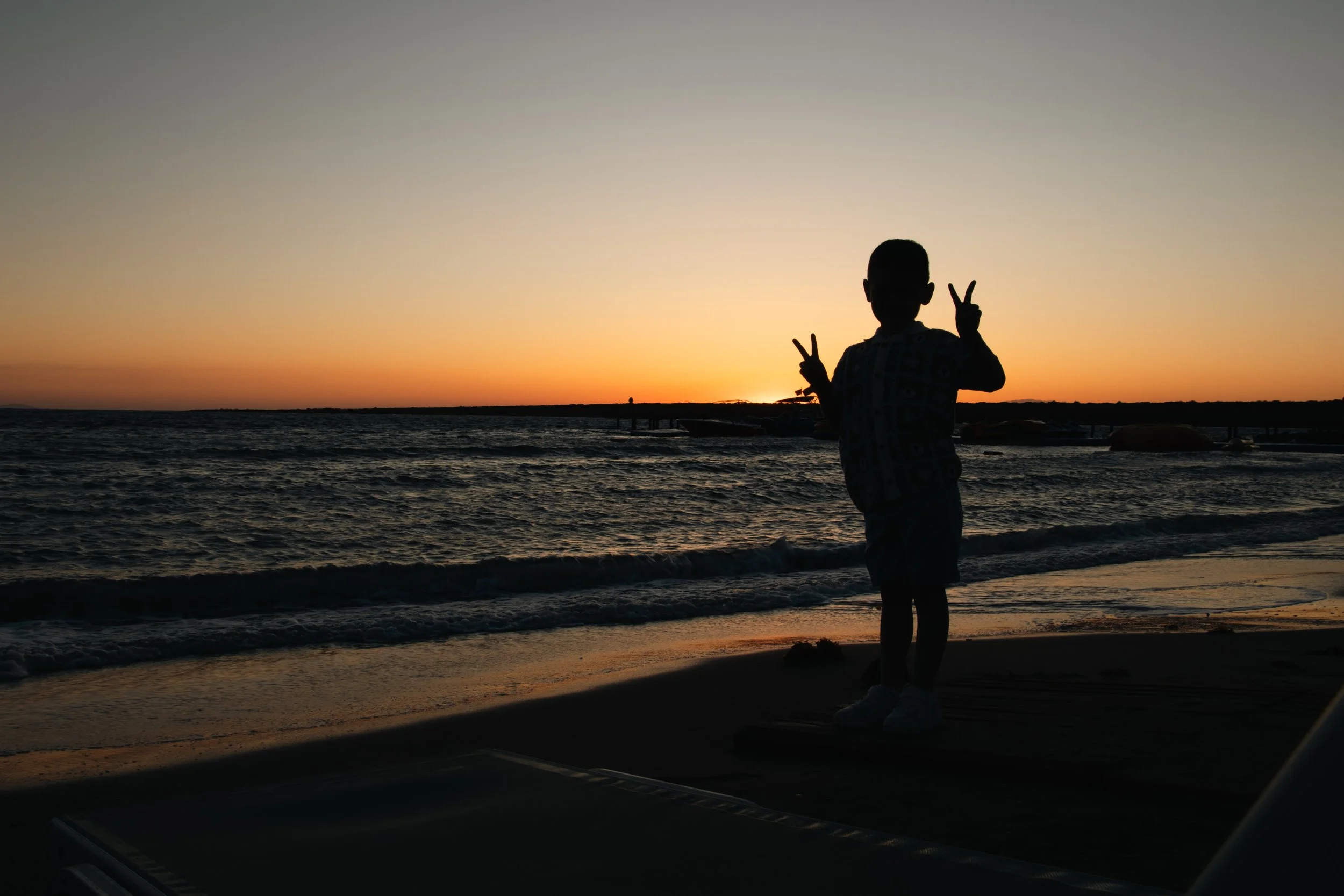 Silhouette of a boy making peace signs with both hands on a beach at sunset.