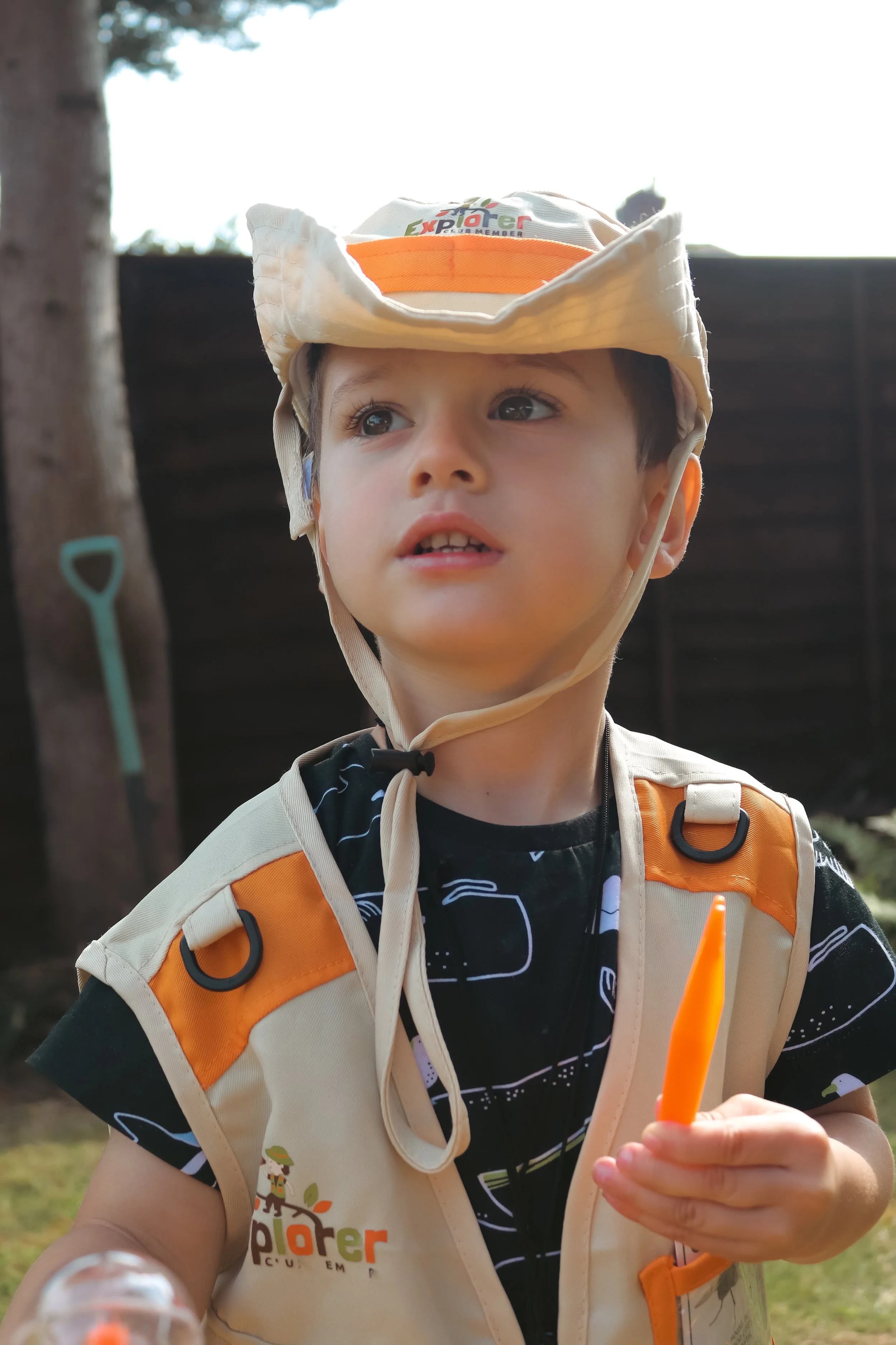 Young boy wearing a ranger hat, orange and beige vest, and holding an orange item outdoors.