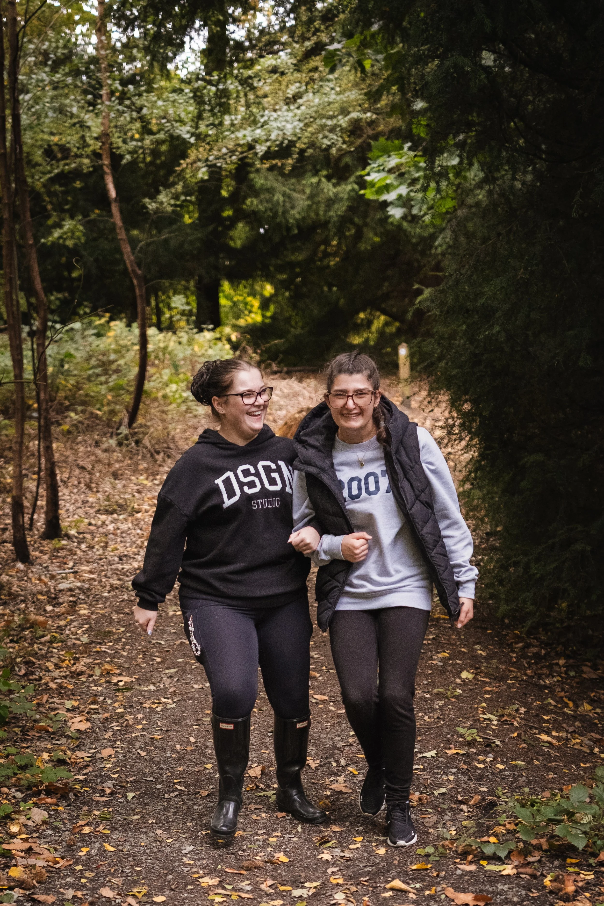 Two young women smiling and walking together on a wooded trail.