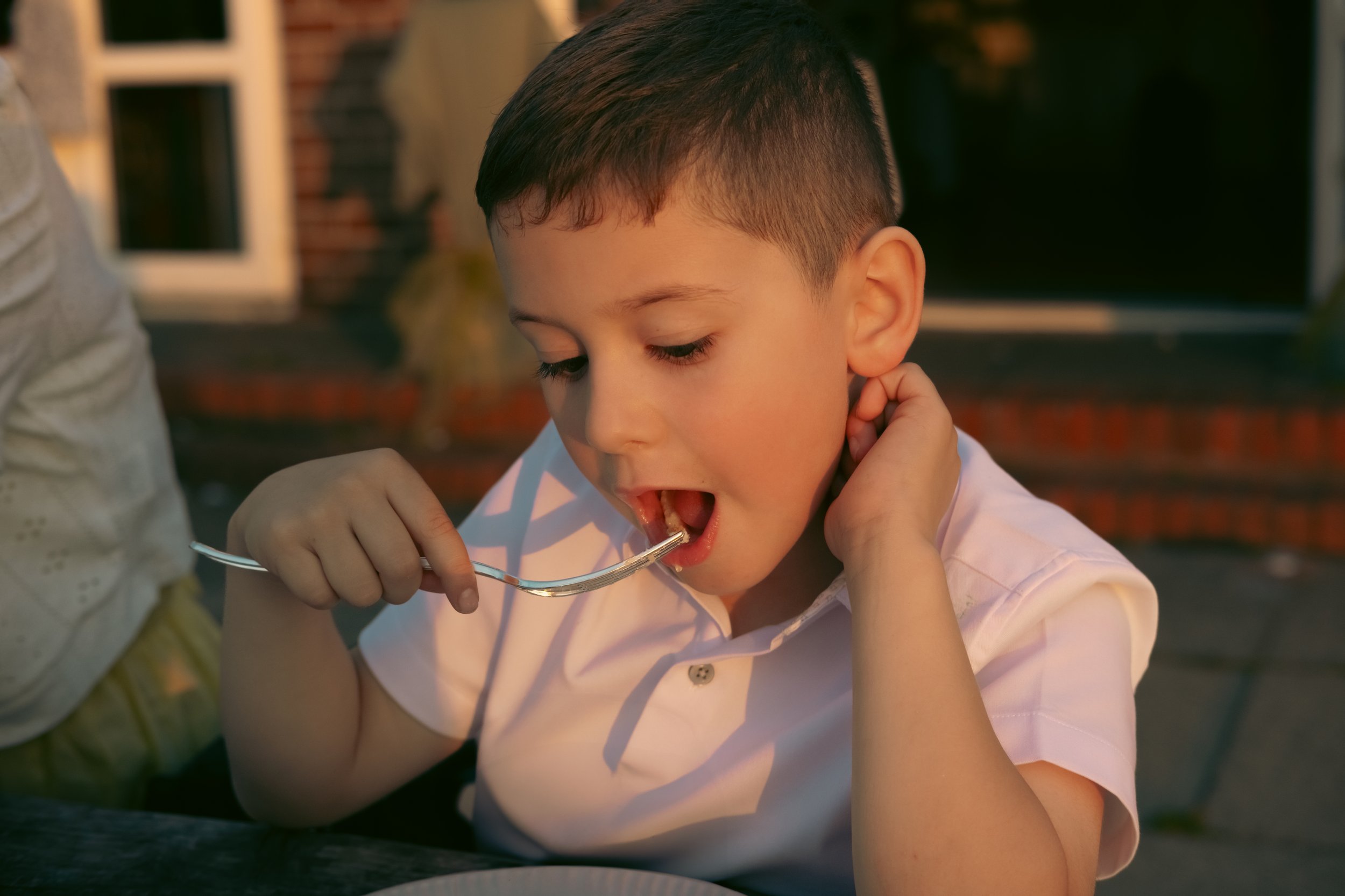 A young boy with short dark hair wearing a light pink shirt eating with a fork outdoors during sunset.