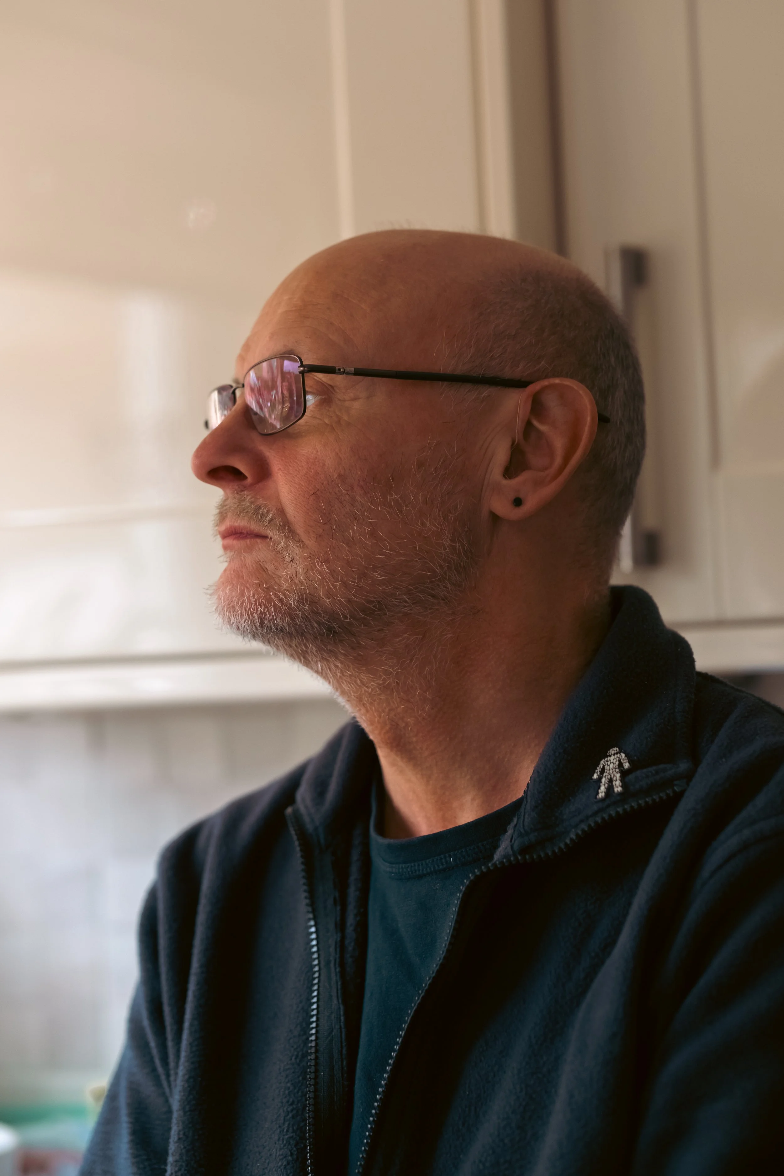 A middle-aged man with glasses, a gray beard, and a bald head looking to the left in an indoor setting with cream-colored cabinets in the background.
