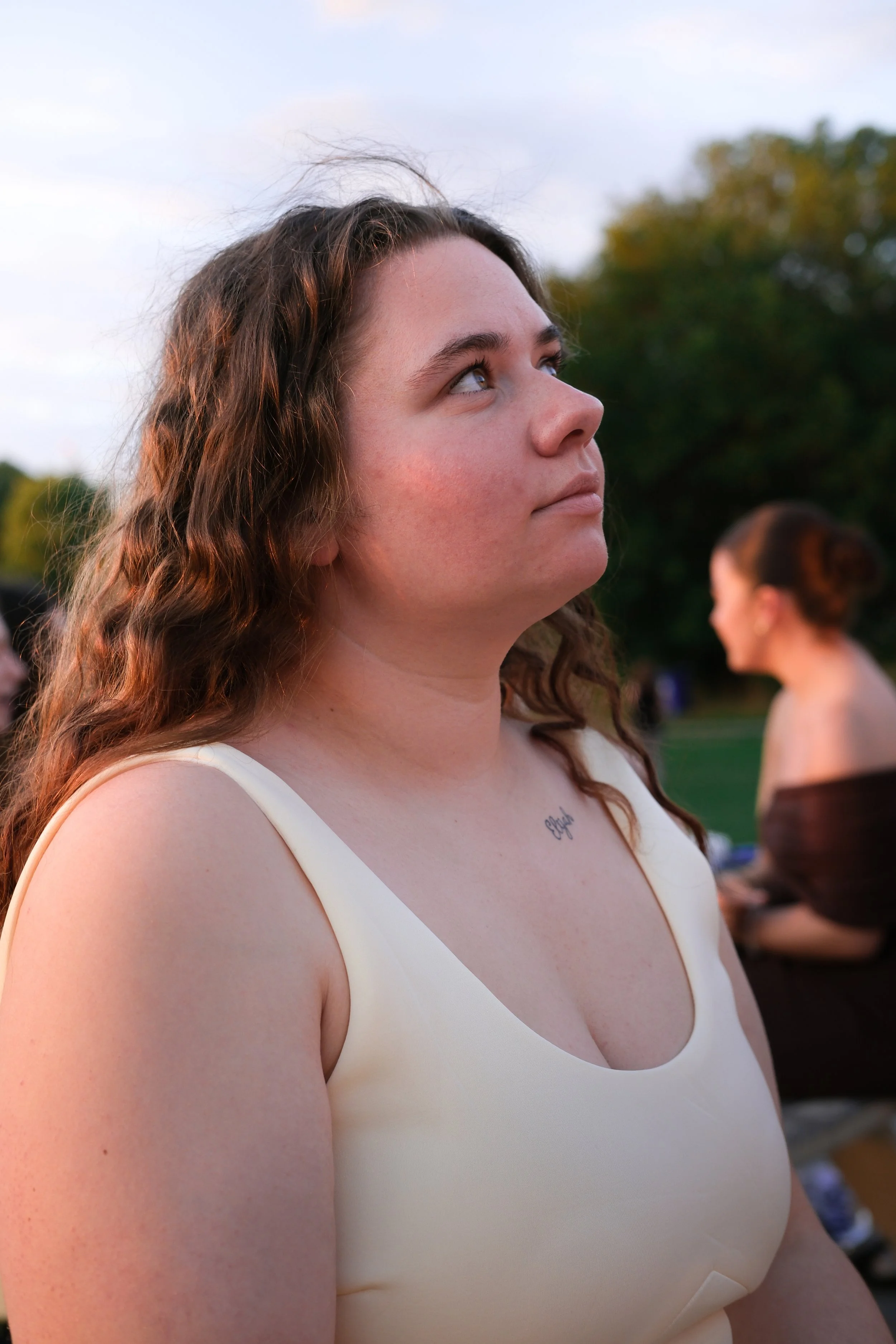 Close-up of a young woman with curly brown hair, wearing a cream-colored sleeveless top, looking thoughtfully to the side outdoors with trees in the background.