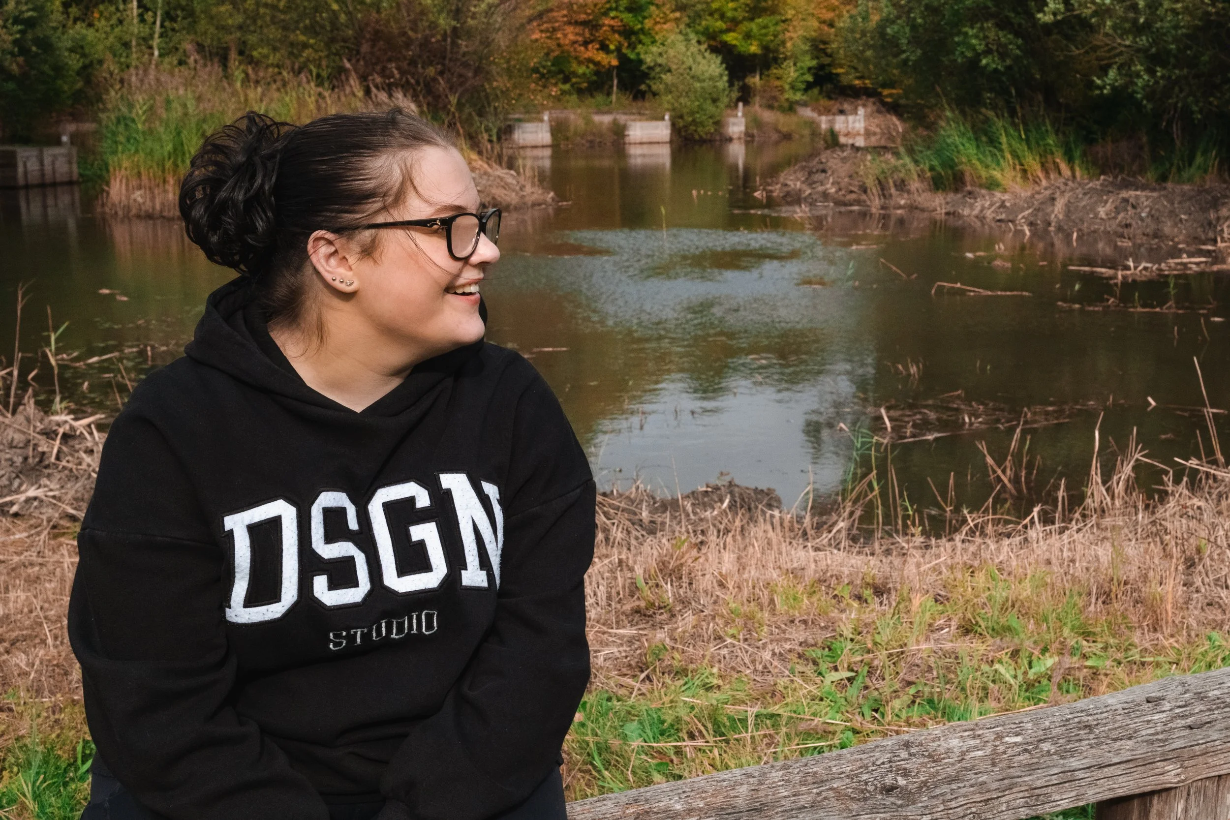 A young woman with glasses wearing a black hoodie, sitting outdoors near a river, smiling and looking to her left, with trees and shrubs in the background.