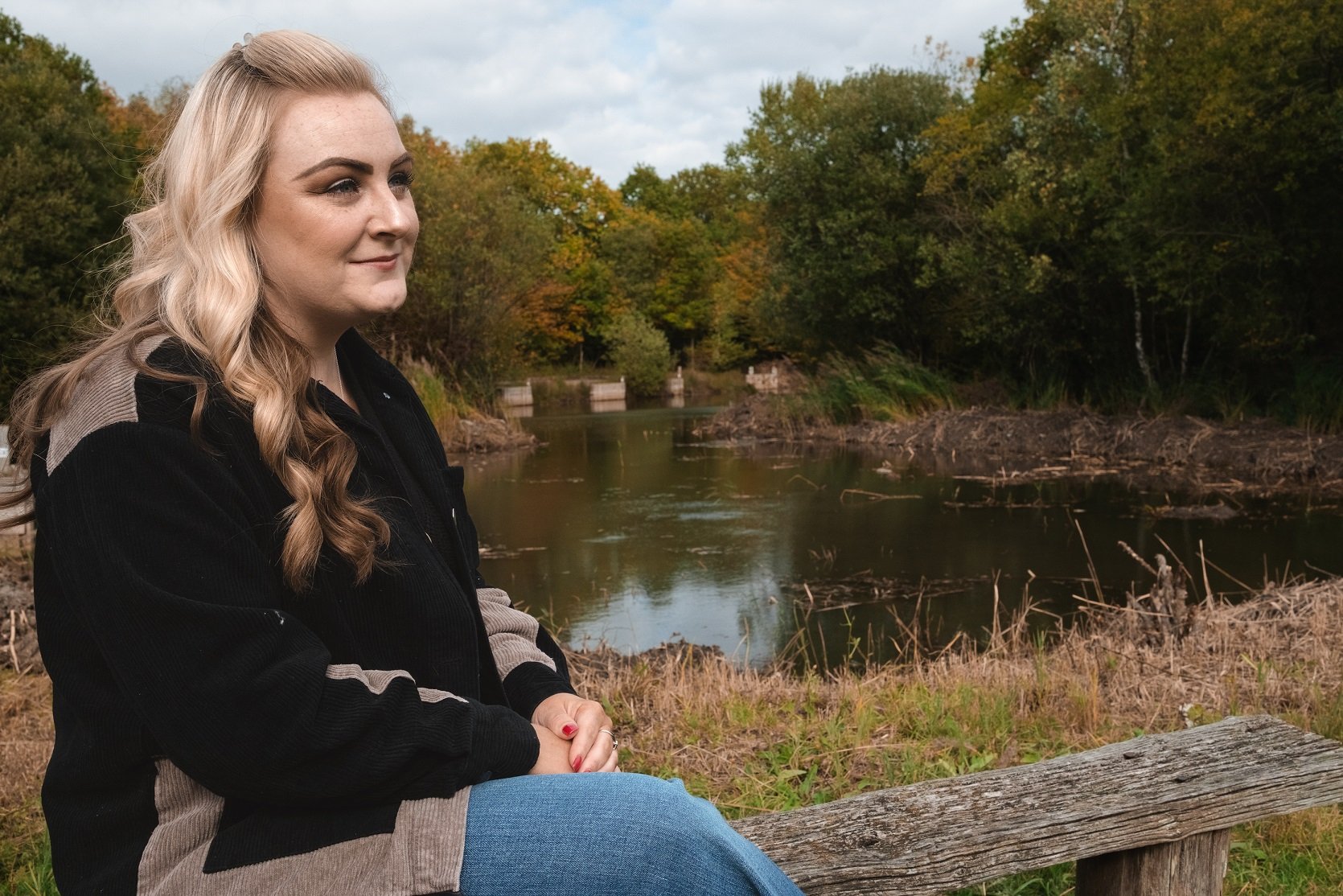 A woman with long blonde hair sitting on a wooden bench outdoors by a river, with trees showing fall colors in the background.