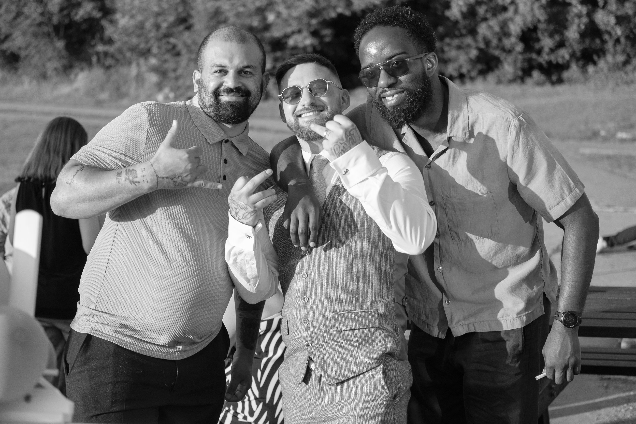 Three men smiling and posing together outdoors, with two of them wearing sunglasses and making hand gestures, in a park-like setting.