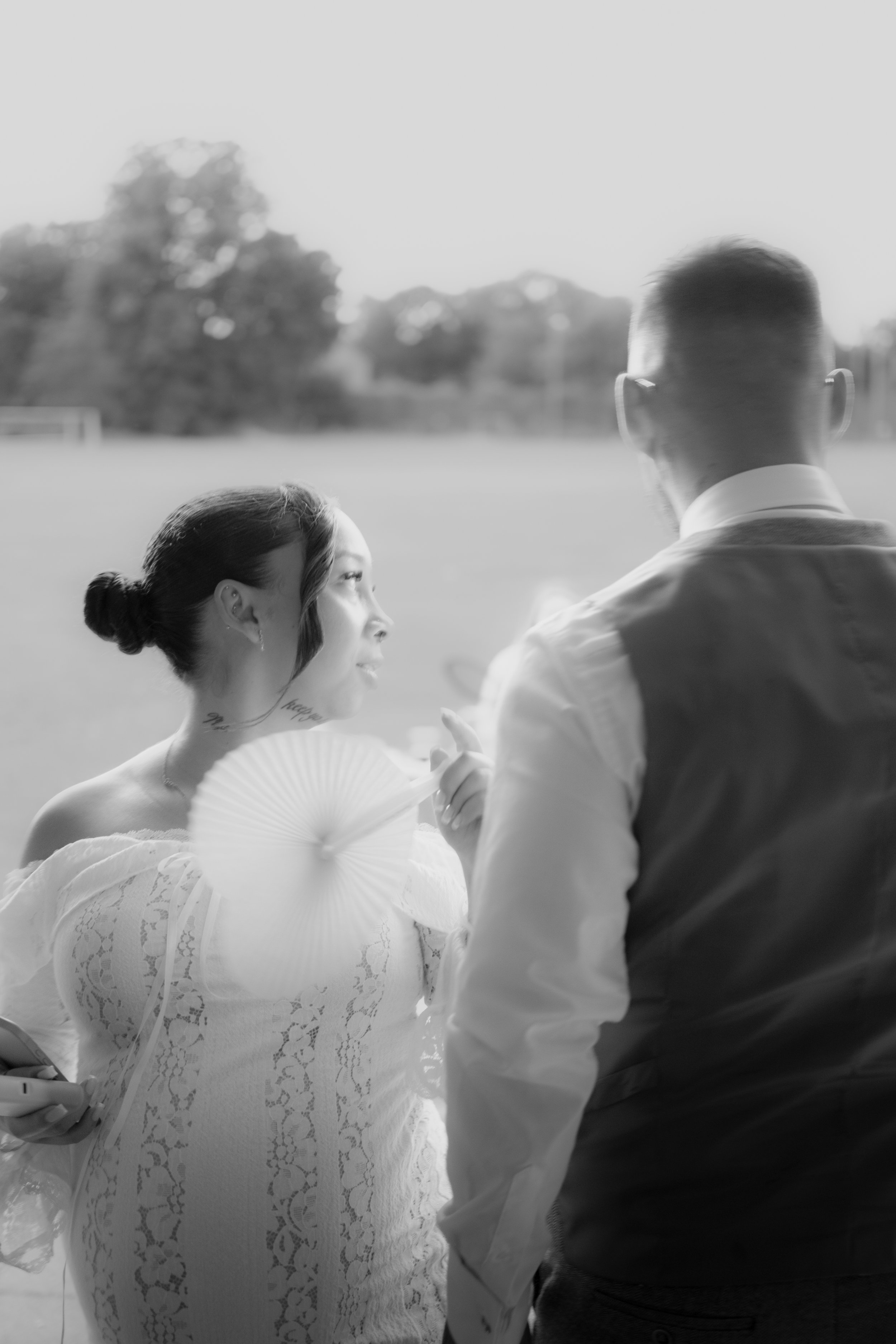 A black and white photo of a woman and a man standing outdoors during sunset. The woman, wearing a lace off-shoulder dress and holding a paper fan, is looking at the man. The man, wearing glasses, a vest, and a long-sleeved shirt, is facing her. The 