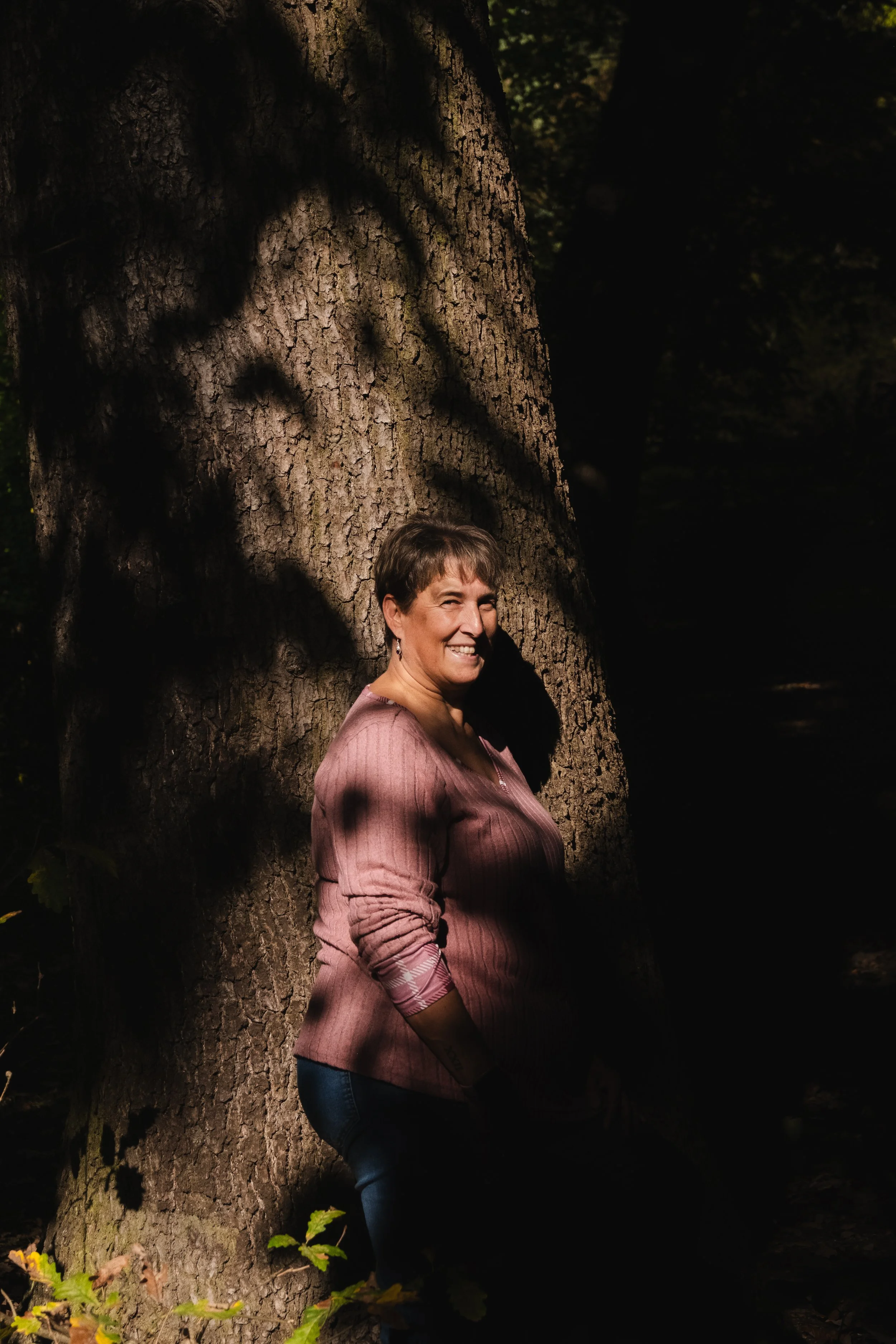 A woman standing outdoors in a forest, smiling, with sunlight casting shadows on her face and a large tree trunk behind her.