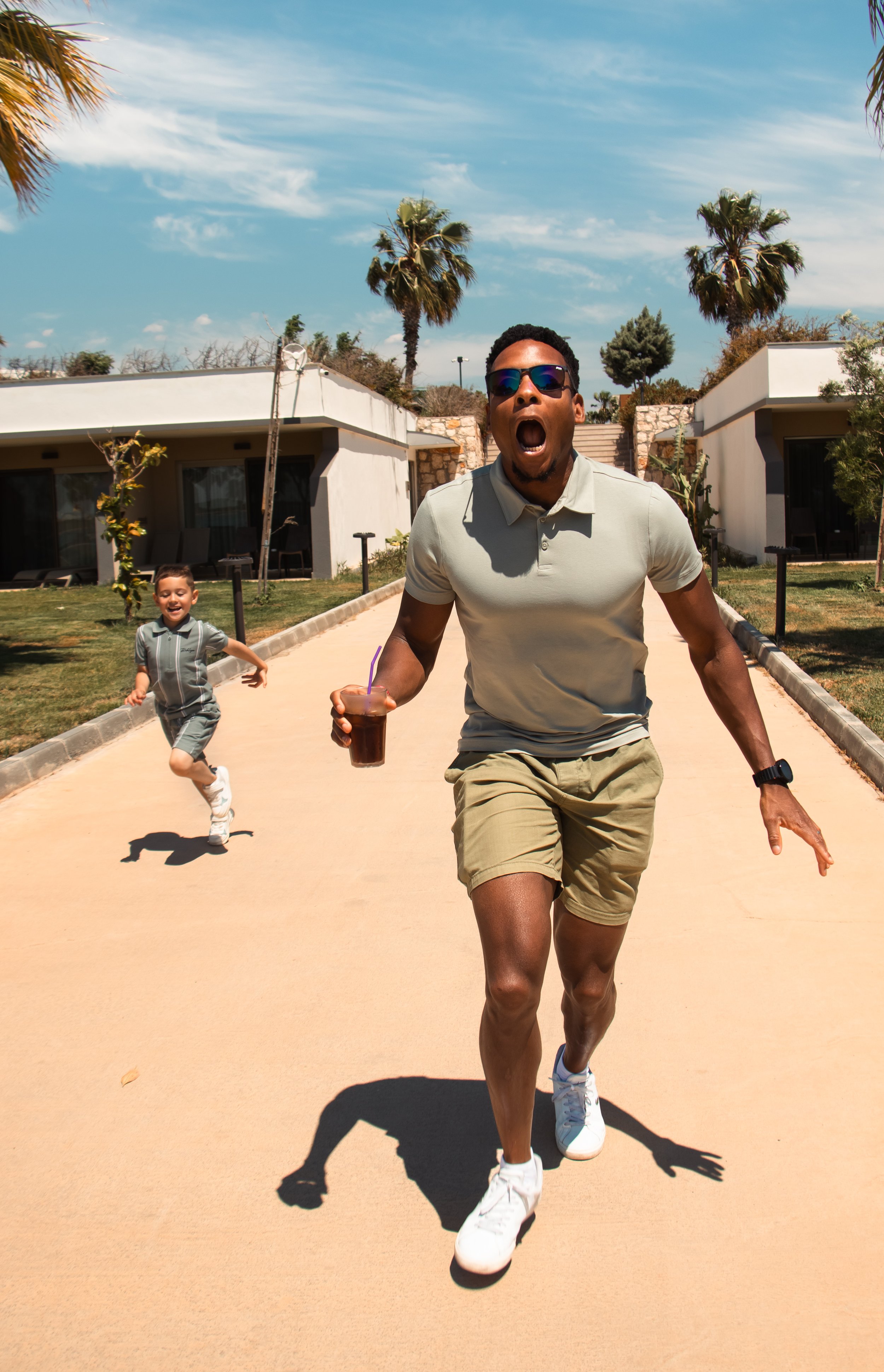 A man and a young boy running outdoors on a sunny day. The man is wearing sunglasses, a light-colored polo shirt, khaki shorts, and white sneakers, holding a drink in his right hand. The boy is dressed in a gray outfit with white shoes, smiling and c
