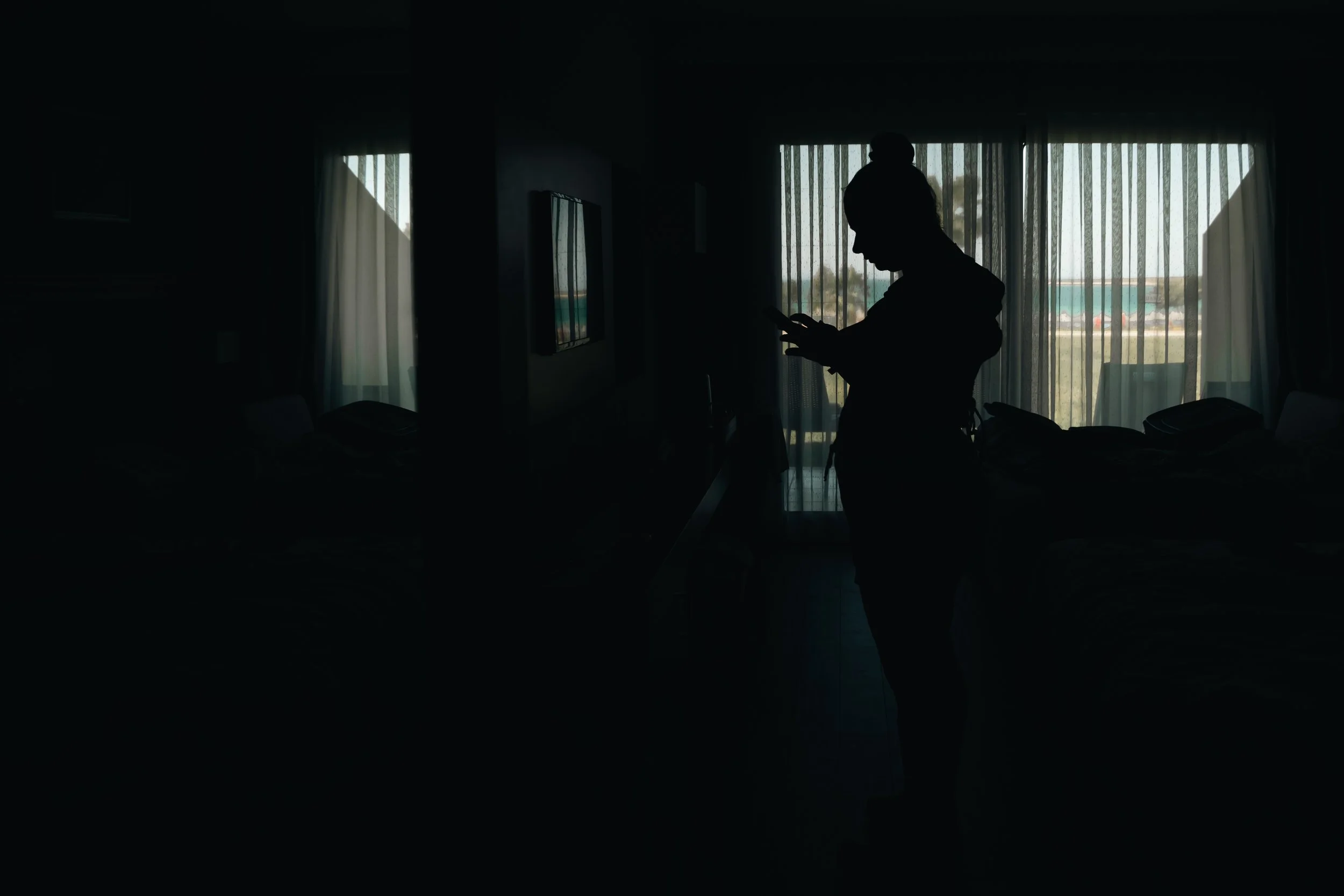 Silhouette of a woman standing inside a dimly lit hotel room, looking at her phone with balcony doors and curtains in the background.