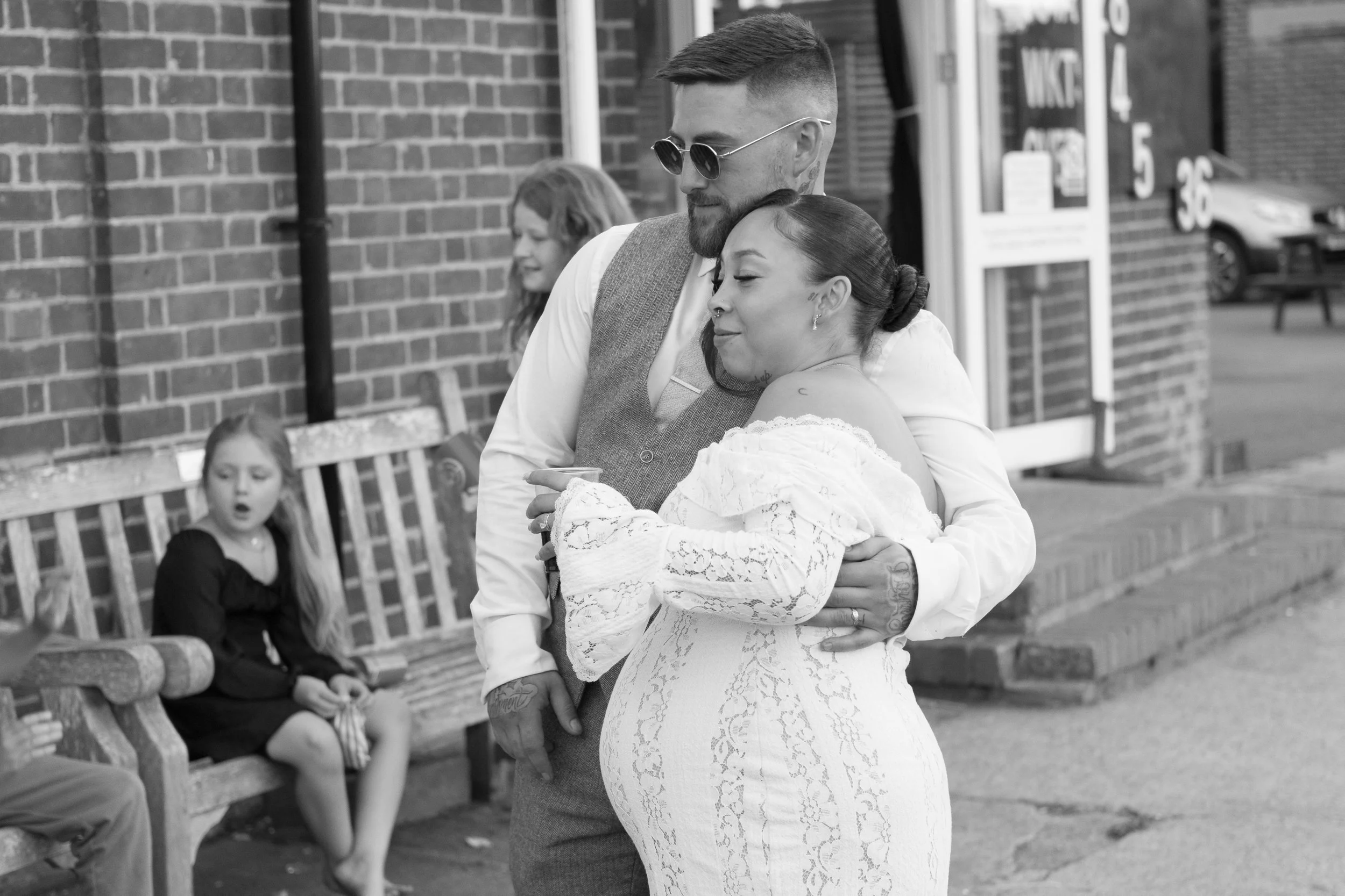 A couple embracing on a sidewalk outside a brick building, with a young girl sitting on a park bench in the background.