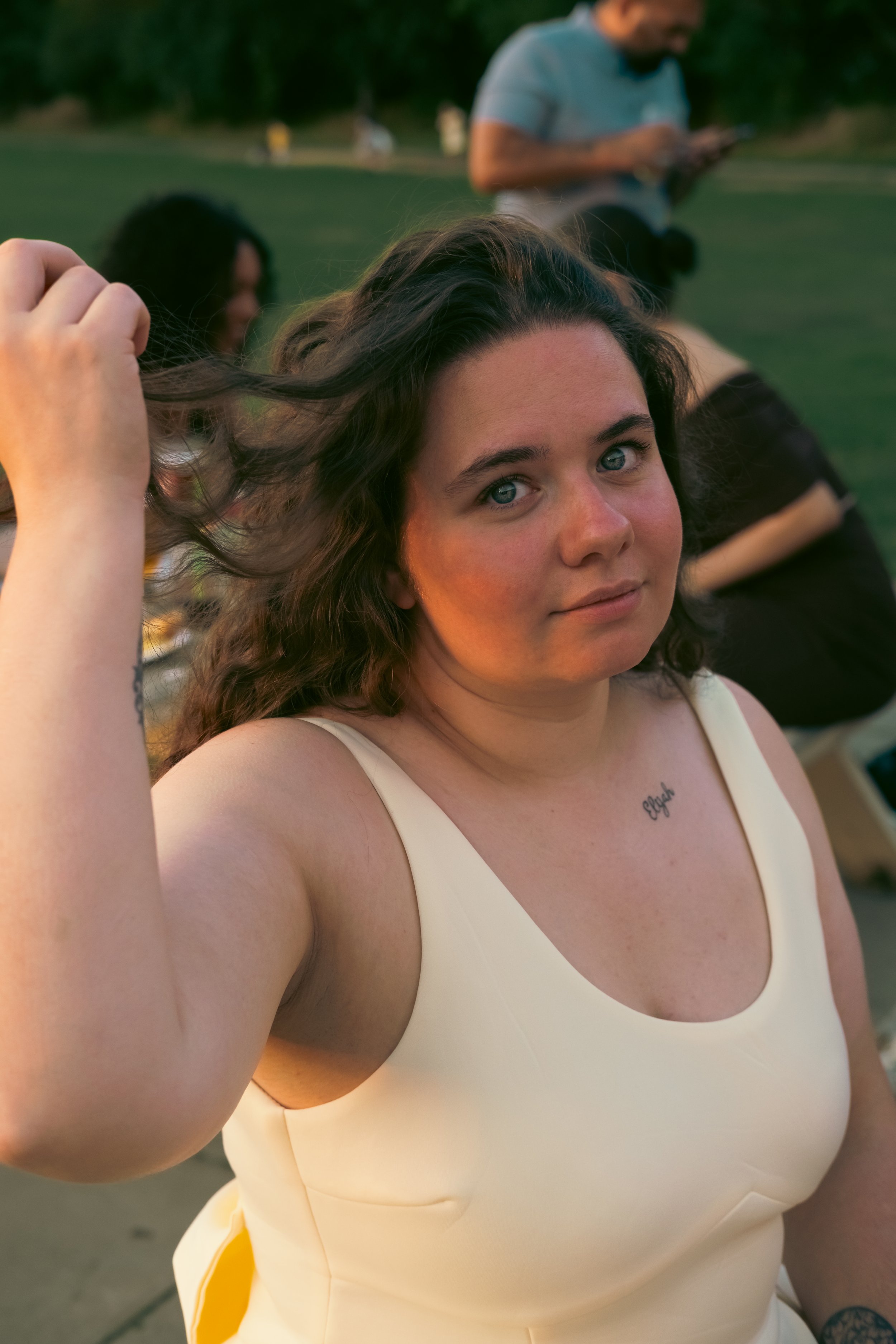 A young woman with curly brown hair and blue eyes taking a selfie outdoors during sunset, with a man in the background looking at his phone and others sitting on the grass.