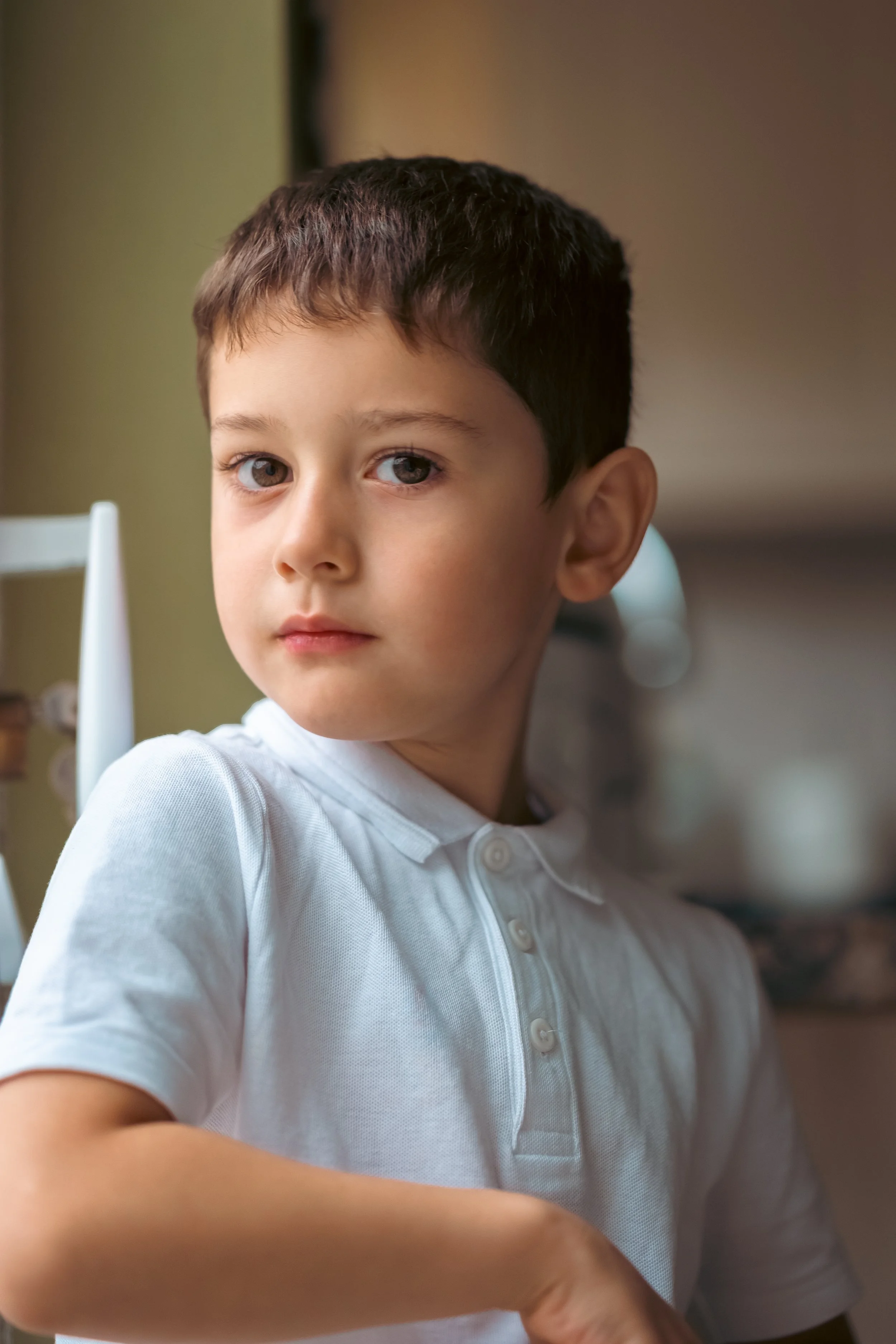A young boy with dark hair and light-colored eyes wearing a white polo shirt, looking slightly over his shoulder in a well-lit room.