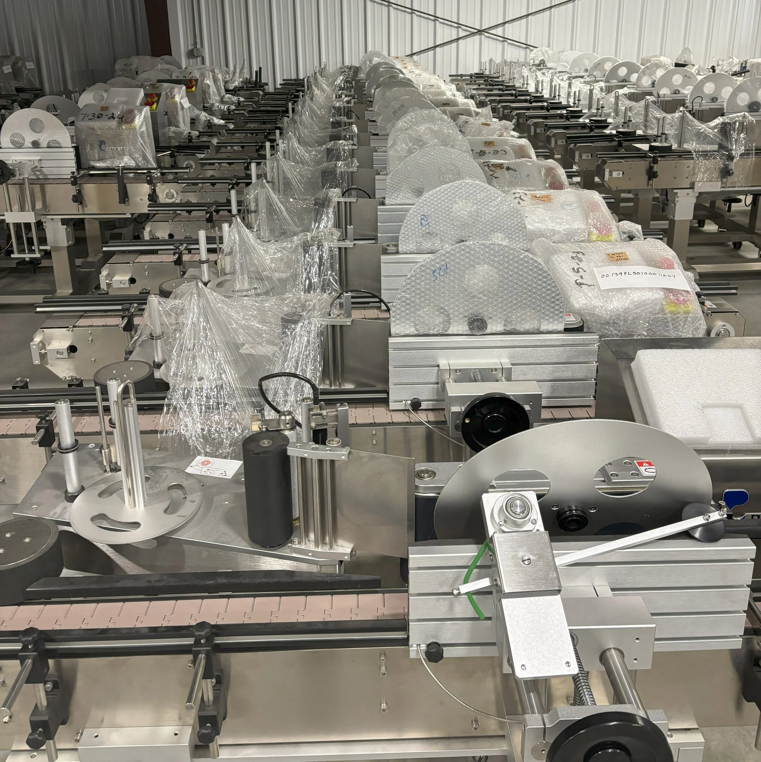 Industrial manufacturing equipment with multiple conveyor belts and components, some wrapped in plastic, in a warehouse setting.