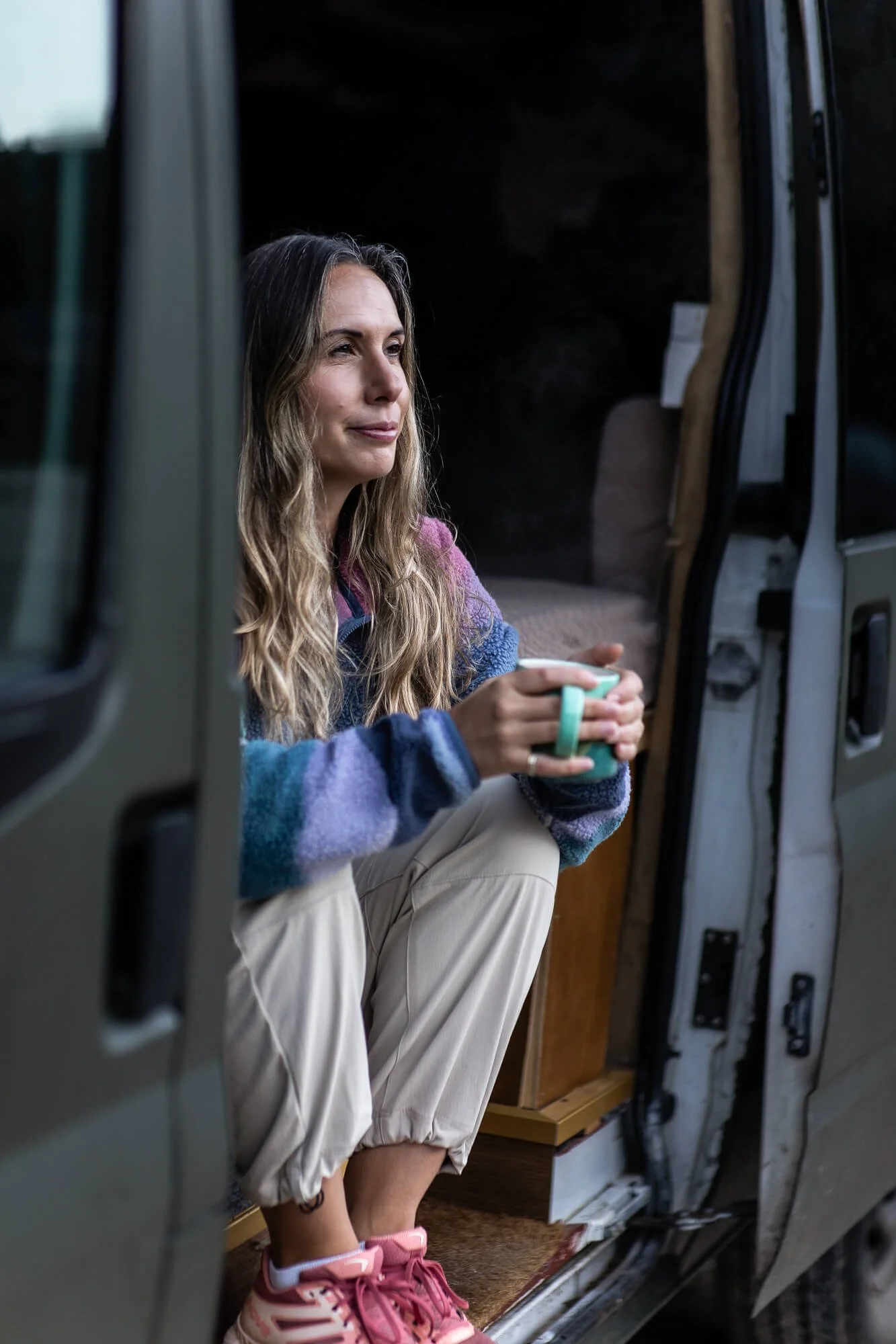 A woman with long wavy hair sits in the doorway of a van, holding a mug and looking thoughtfully into the distance, wearing a colorful sweater, beige pants, and pink sneakers.