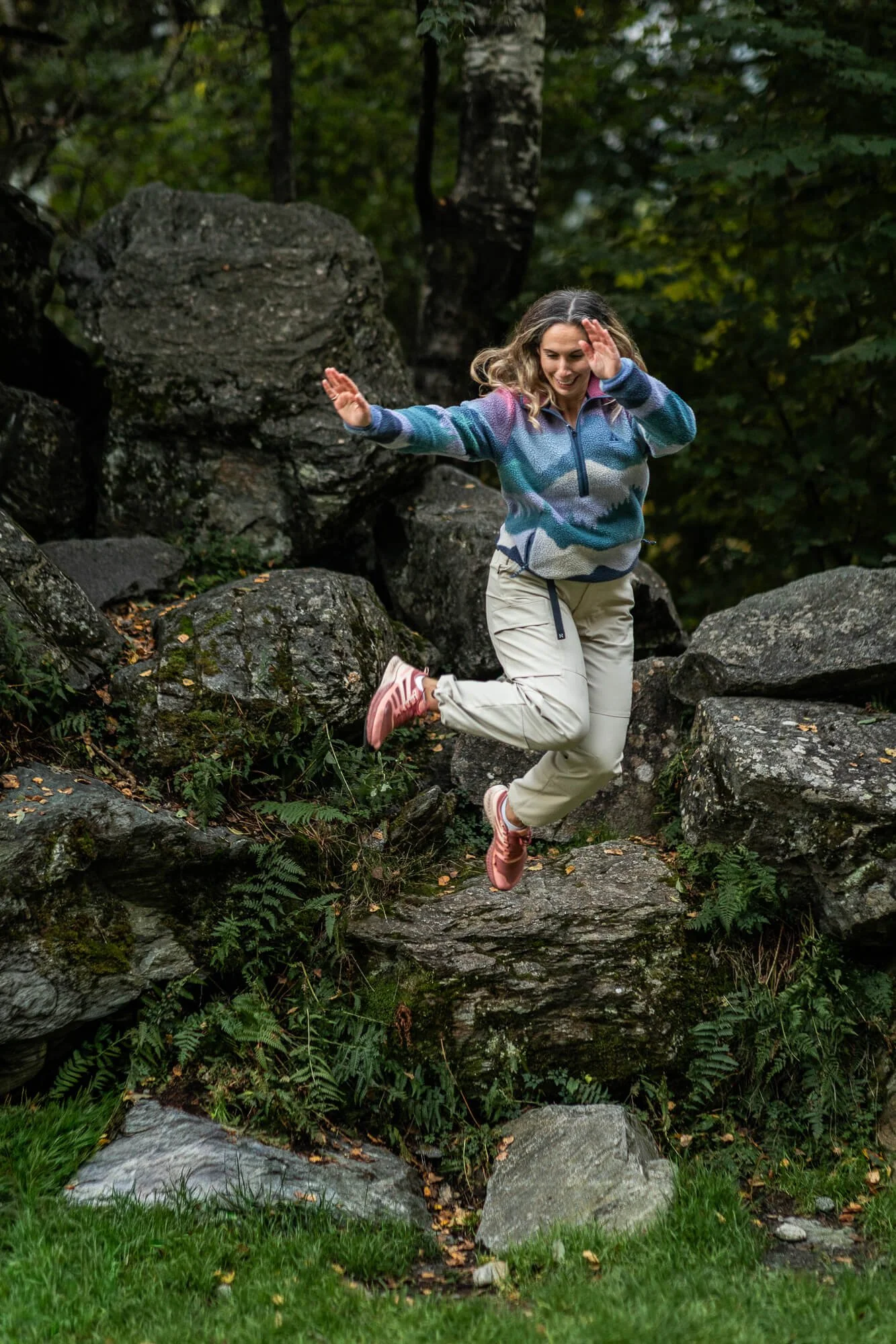 A woman in outdoor gear jumping off rocks in a forested area.