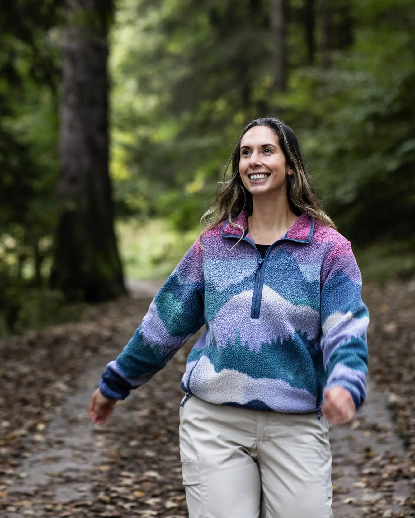 A smiling woman walking in a forest trail, wearing a colorful fleece jacket and khaki pants.