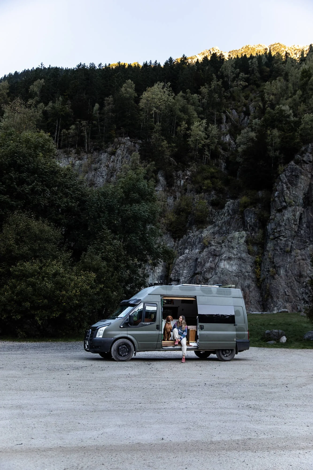 A woman and a dog sitting on the step of a parked camper van in front of a mountainous landscape with trees and rocks.