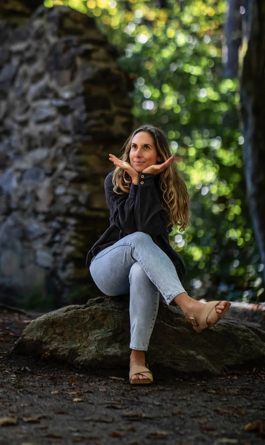 A woman sitting on a rock in a forest, with greenery and sunlight in the background, resting her chin on her hands and smiling creatively.