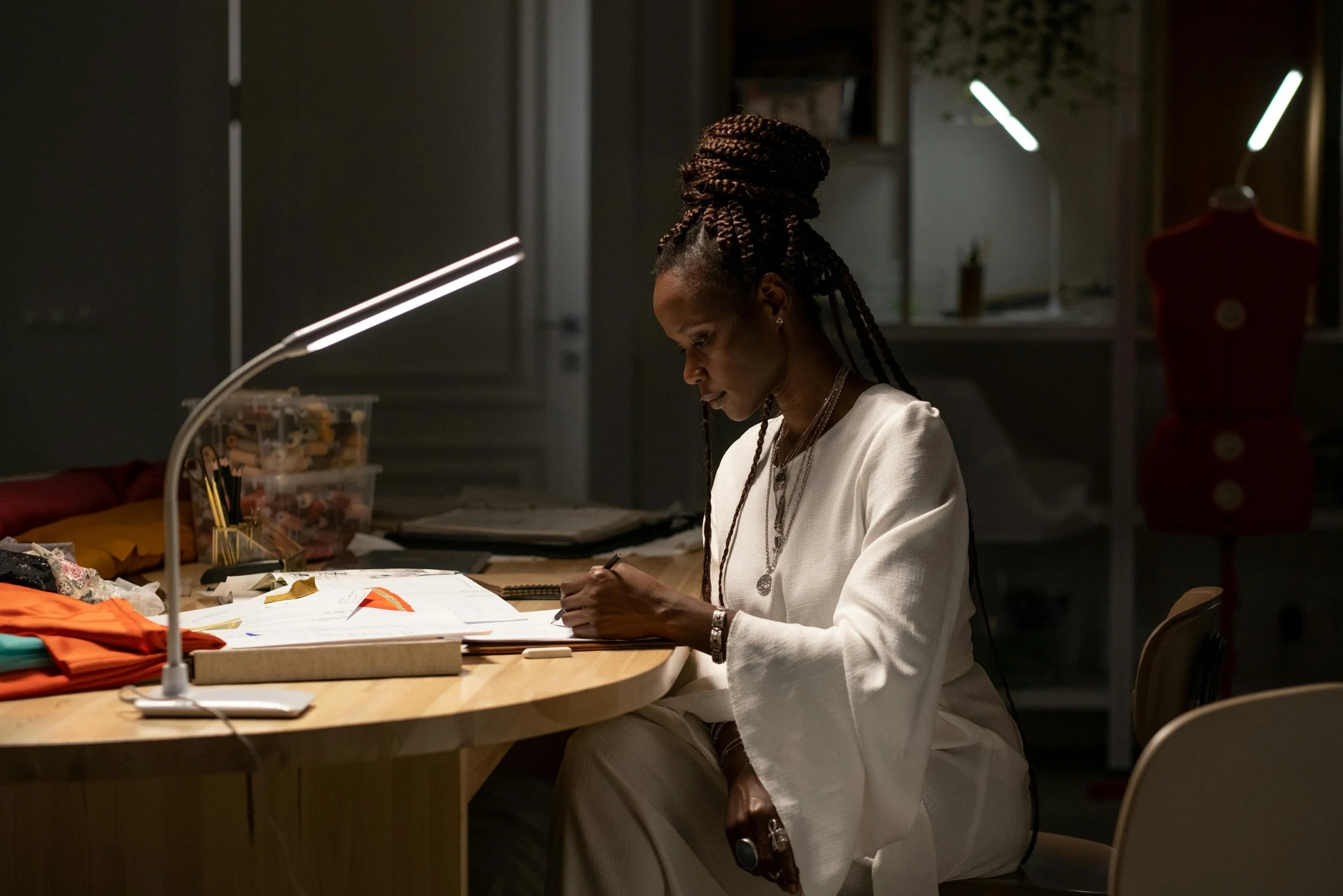 This image shows a woman working at a desk late at night reviewing documents. She has dark braided hair and is holding a pencil. She is sketching something onto paper.