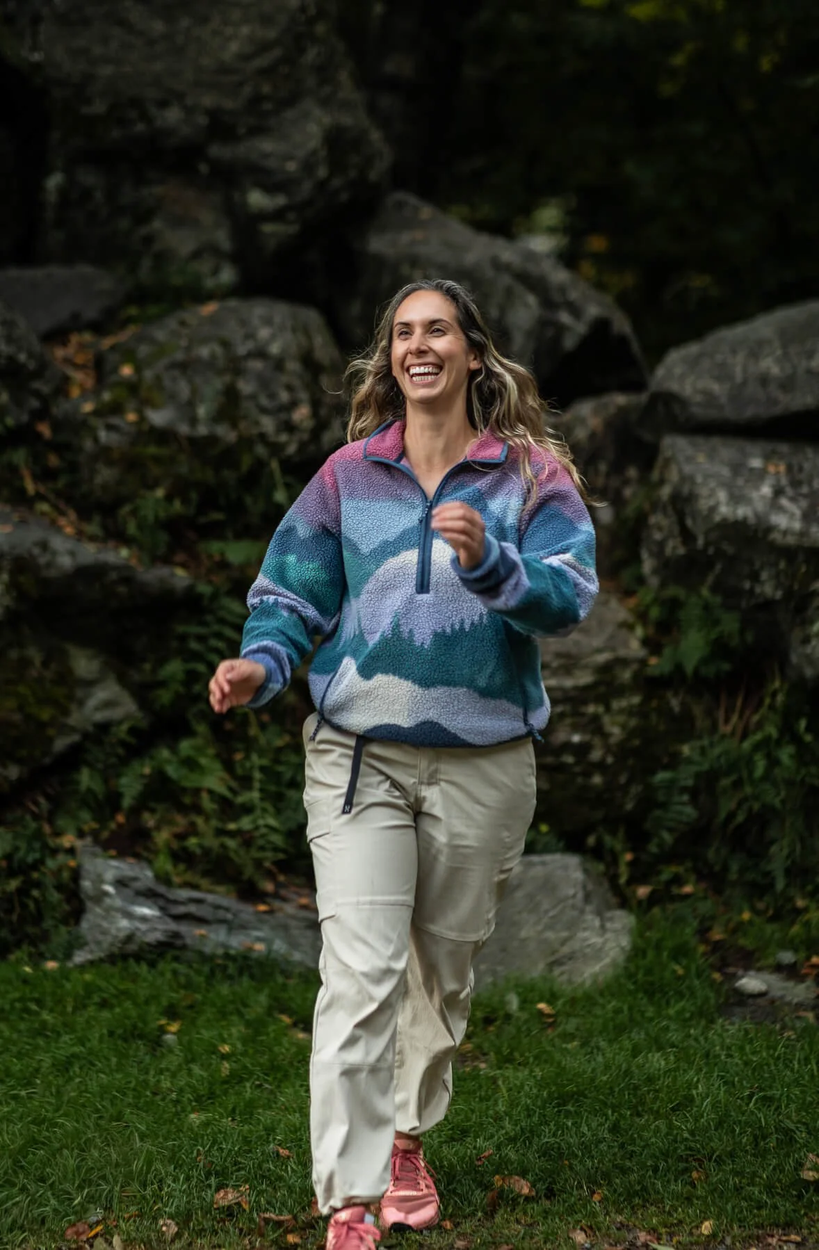 A woman with long, wavy hair smiling while jogging outdoors in a forested area, wearing a colorful fleece jacket, beige pants, and pink running shoes.