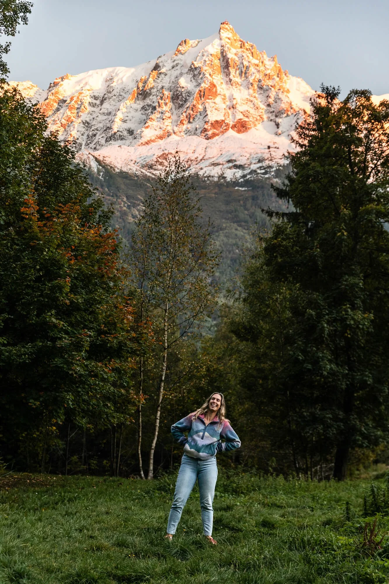 A woman wearing a colorful jacket and jeans standing on a grassy area with trees, smiling, with a snow-capped mountain in the background during sunset.