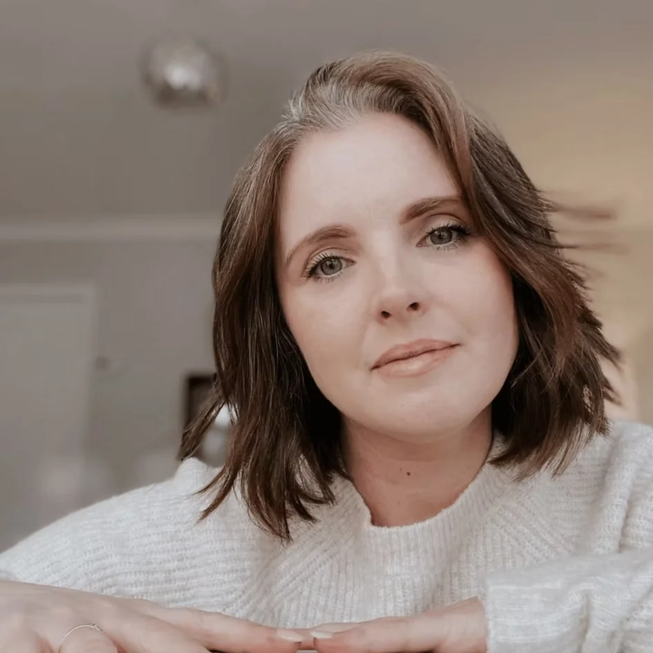 A woman with shoulder-length brown hair and light eyes, wearing a cream-colored sweater, resting her arms on a surface and looking into the camera in a well-lit indoor setting.