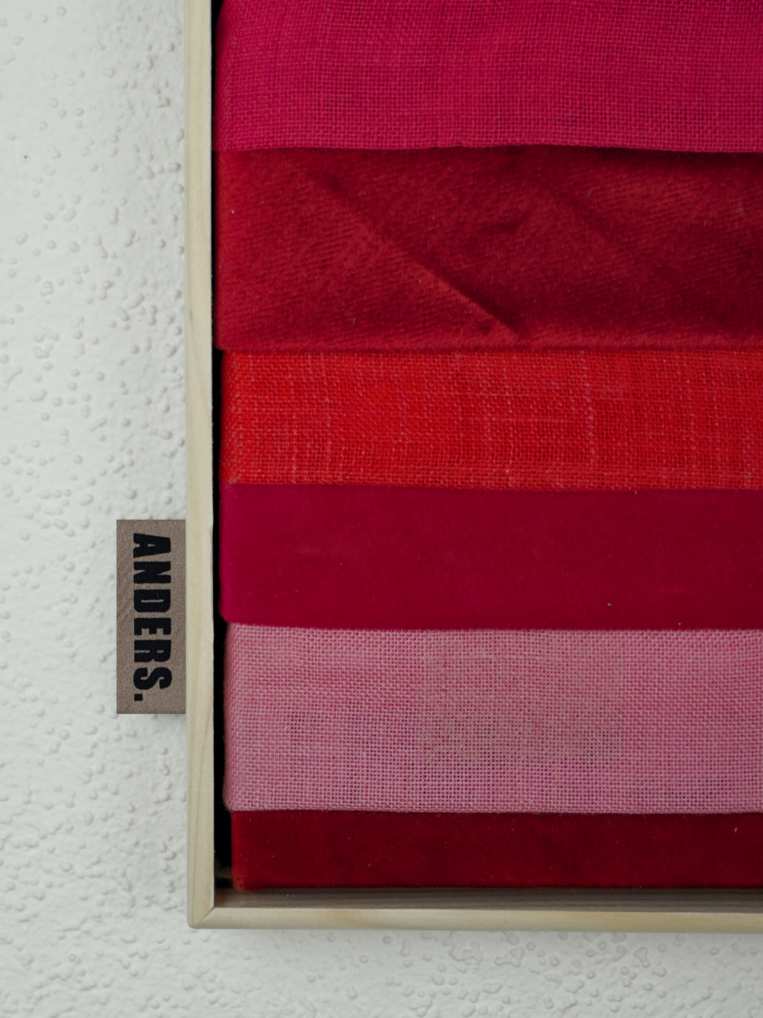 Verschiedene Stoffproben in Rottönen, präsentiert in einer Holzschatulle, neben einer Etikette mit der Aufschrift 'ANDERS'.