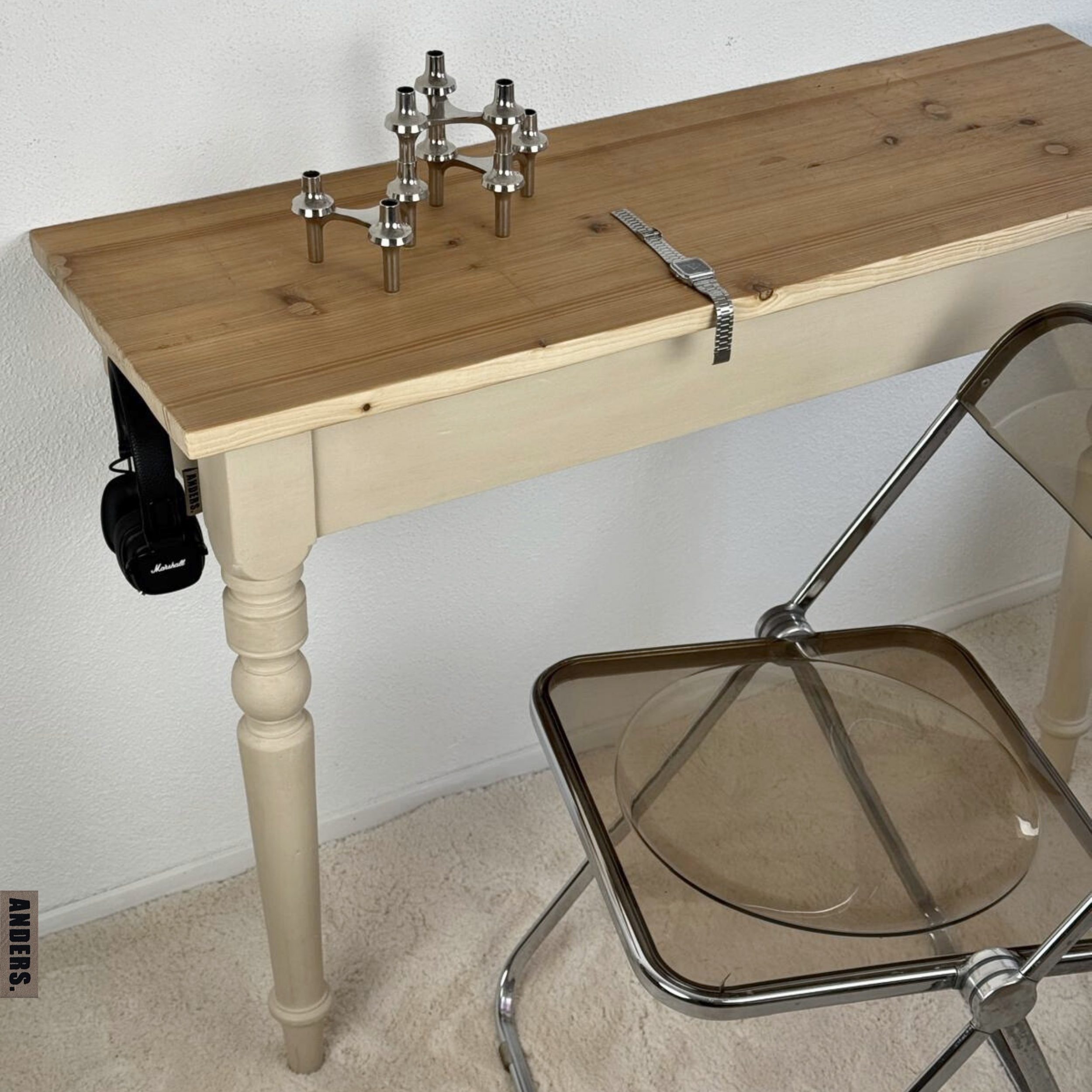 Holztisch mit Metallkugeln und Uhrenarmband, begleitet von einem transparenten Stuhl, im Raum mit weißen Wänden und Teppichboden.