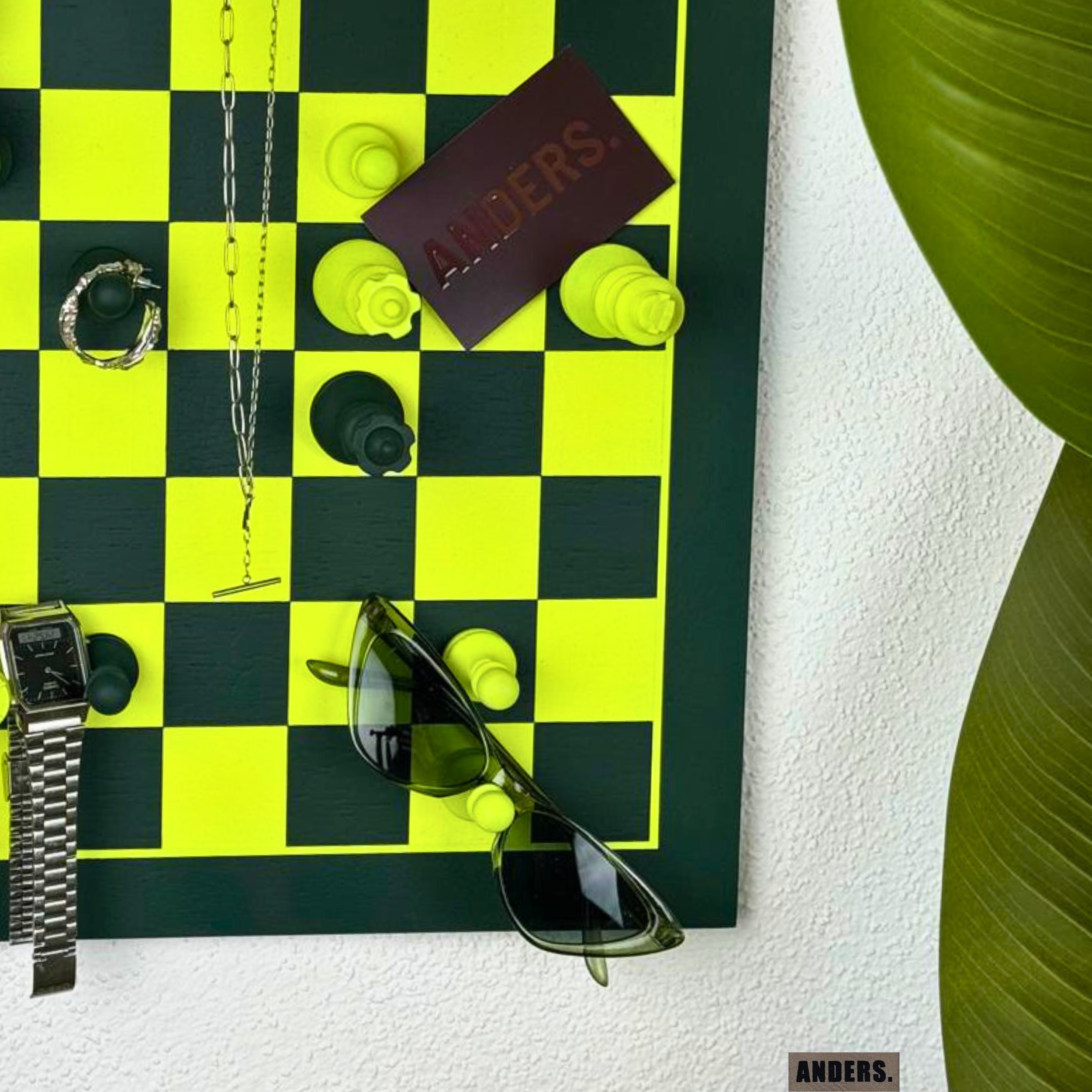 Ein Schachbrett mit Schachfiguren, eine Uhr, eine Sonnenbrille, eine Halskette, ein Notizbuch mit der Aufschrift 'ANDERS' und ein Armband, alles auf einer weißen Oberfläche neben einer grünen Pflanze.