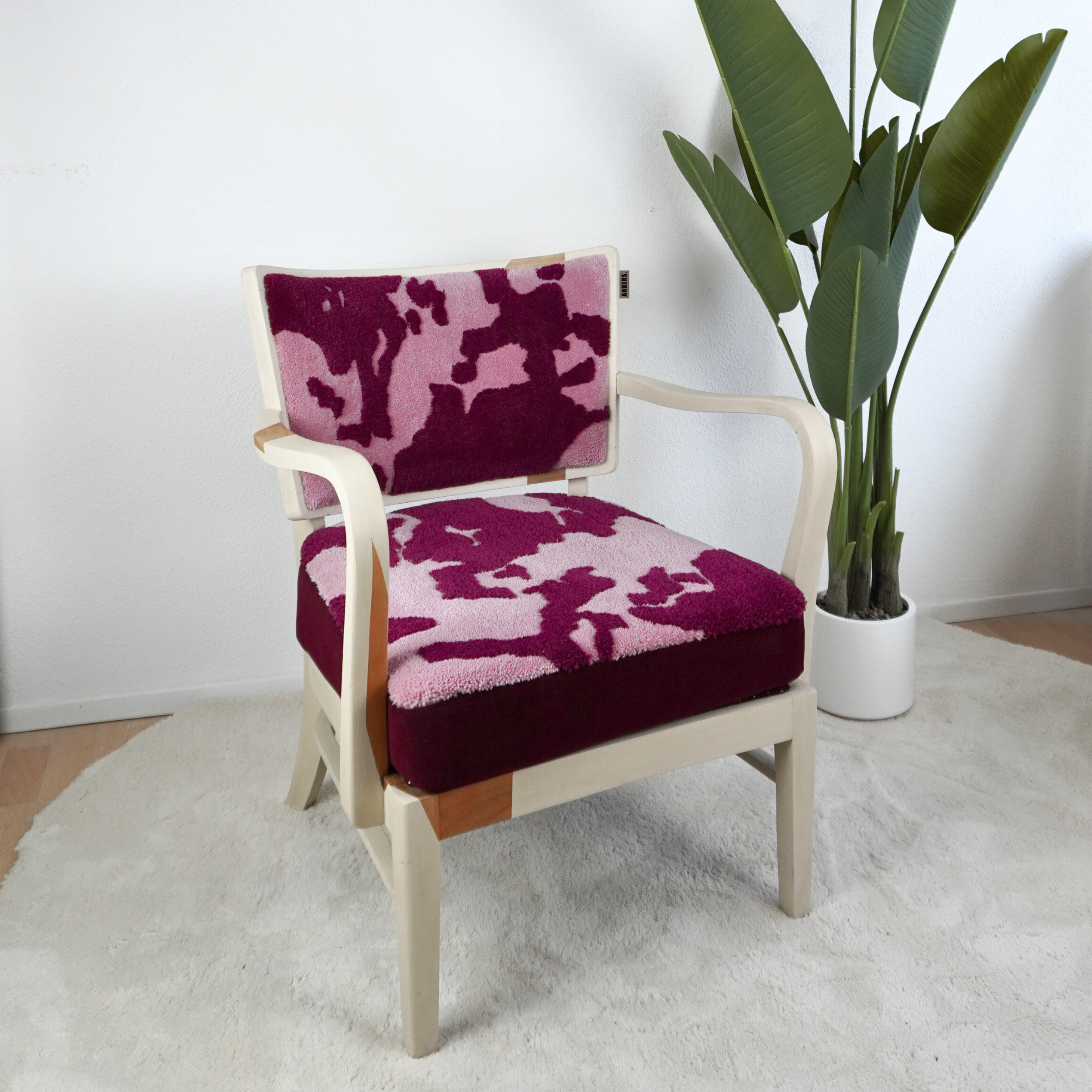 Ein Sessel mit einer dreifarbigen, gemusterten Polsterung in Pink, Rot und Dunkelrot neben einer großen Topfpflanze in einem weißen Raum.