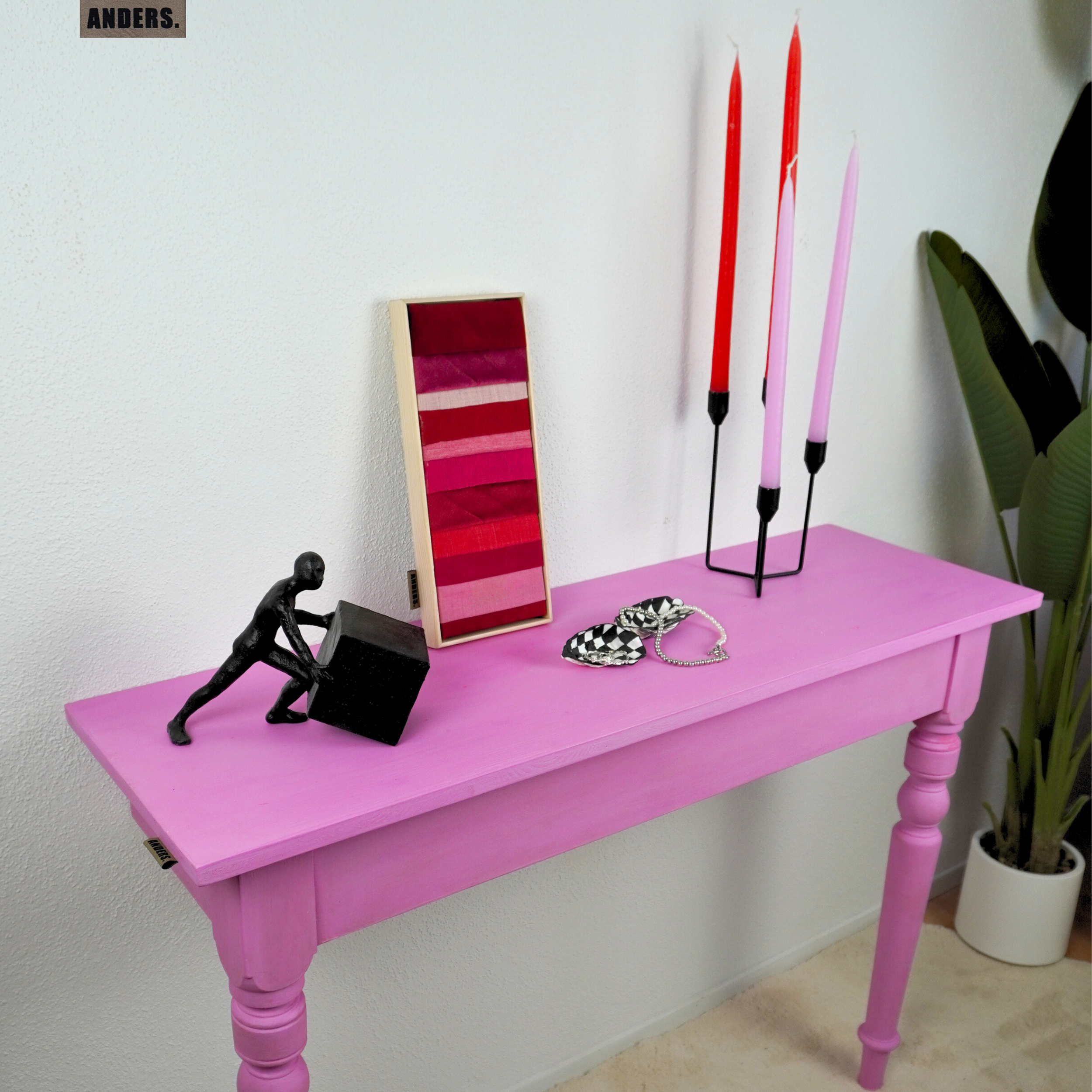 Ein pinker Tisch mit verschiedenen Dekorationsgegenständen, darunter eine schwarze Figur, ein Farbmuster, eine Kette, und vier Kerzen in unterschiedlichen Farben, vor einer weißen Wand