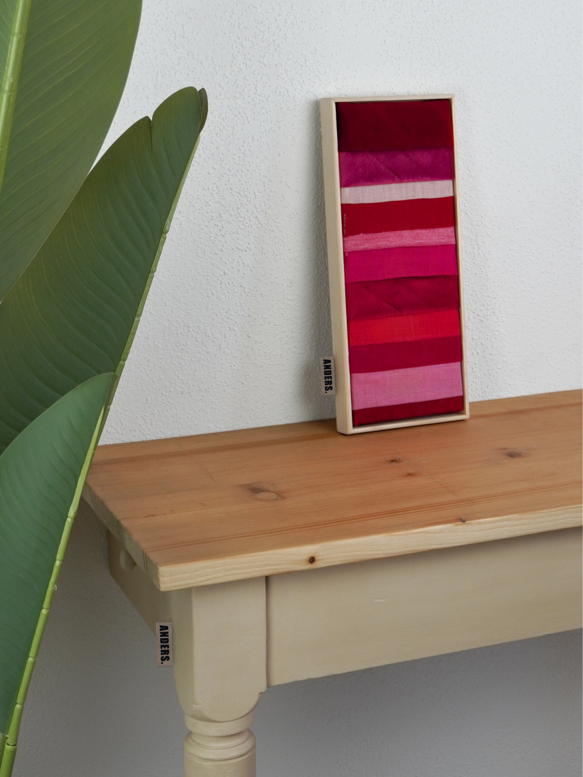 Eine Holzwand mit einem schmalen, vertikal hängenden Rahmen, der Farben in verschiedenen Rottönen enthält, neben einem großen grünen Blatt einer Pflanze.