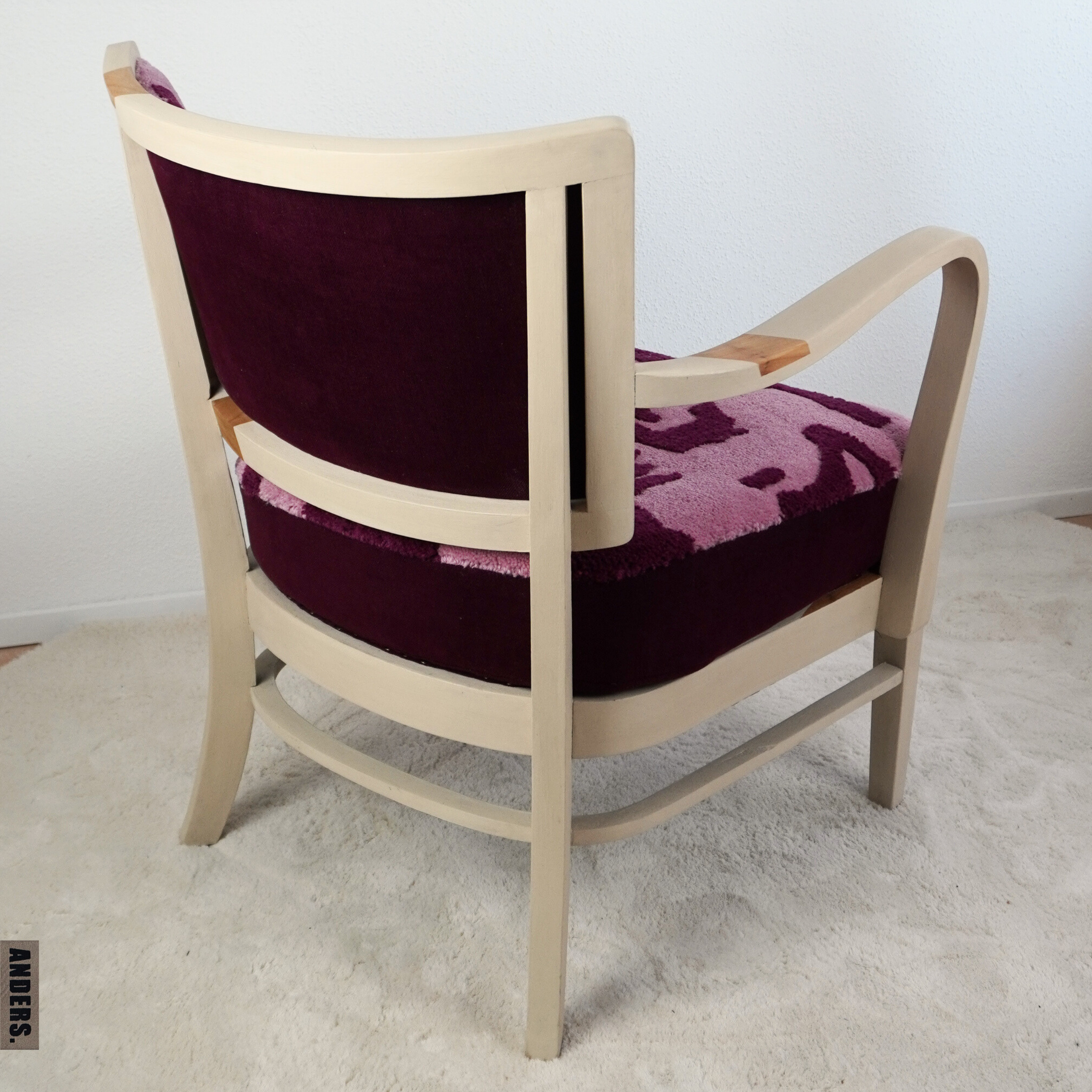 Ein Stuhl mit cremefarbenem Holzrahmen und dunkelrotem samtartigem Polster, der an einem weißen Wand steht.